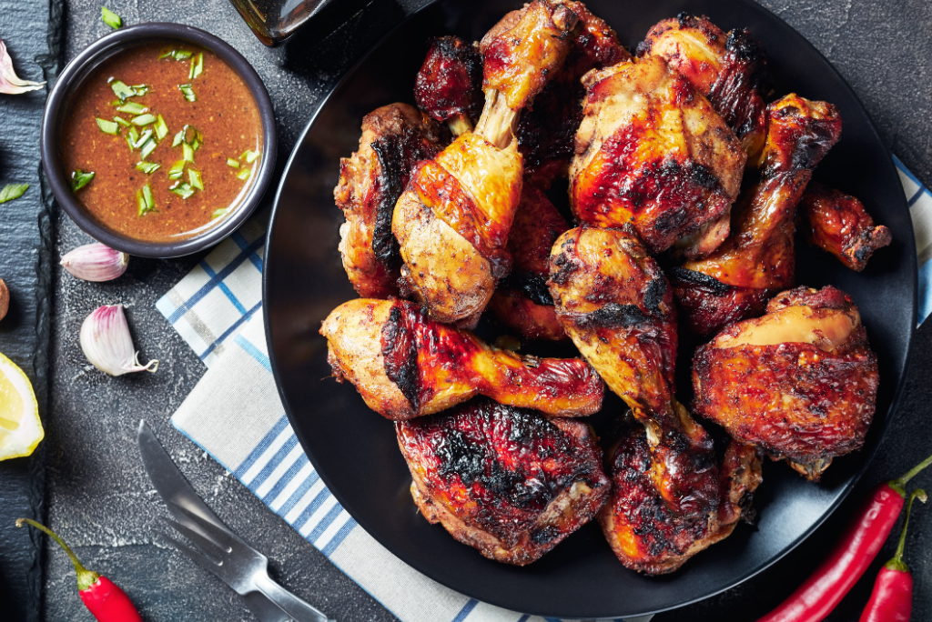 Plate of grilled chicken drumsticks with barbecue sauce, accompanied by a small bowl of sauce garnished with green onions, on a dark surface with garlic, lemon, and red chili peppers nearby.