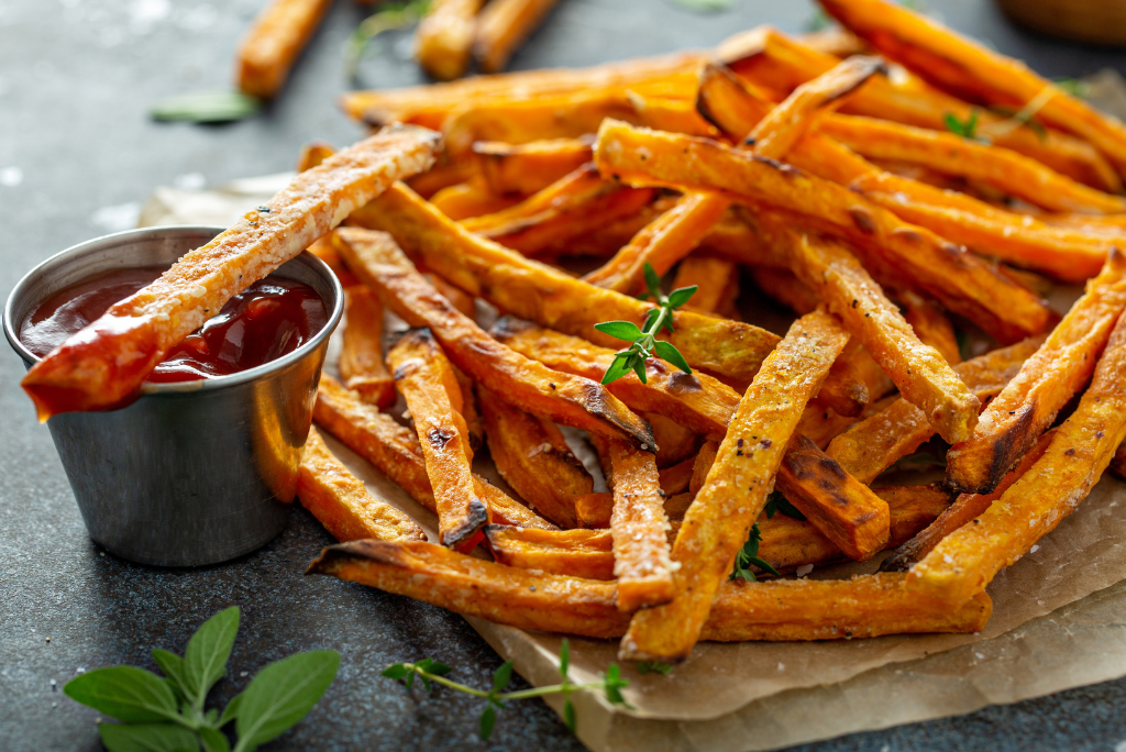 A plate of crispy sweet potato fries garnished with herbs, served with a small cup of ketchup for dipping.