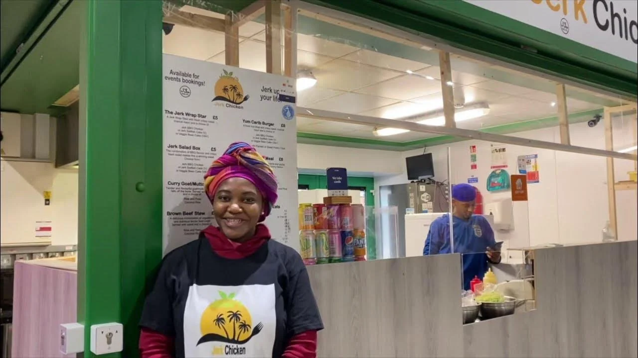 A smiling woman wearing a colorful head wrap and black t-shirt with a logo for a chicken restaurant, standing in front of a food stall with a menu board behind her. There is another person in the background wearing a blue uniform and cap, preparing food.
