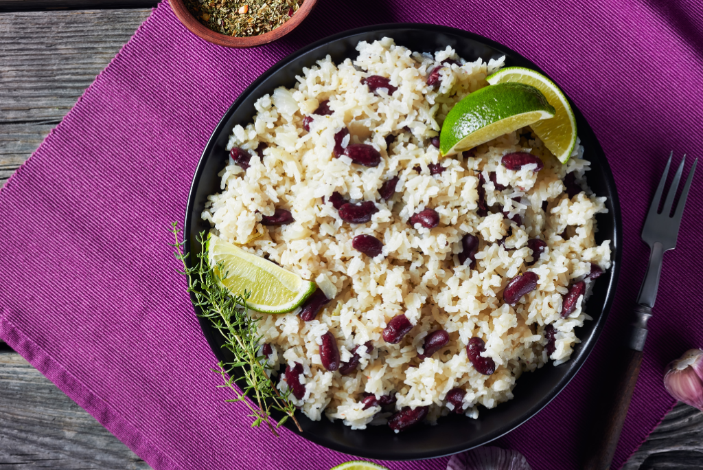 A bowl of rice and kidney beans garnished with lime wedges and a sprig of fresh herb, placed on a purple cloth and wooden table.