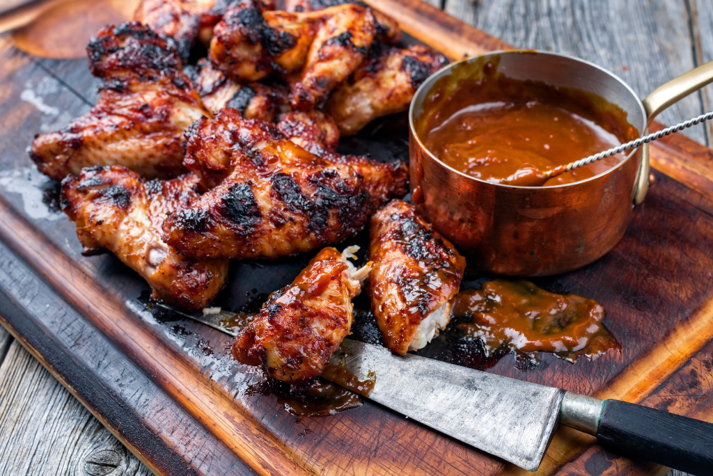 Barbecue chicken wings with sauce on a wooden cutting board, with a copper pot of barbecue sauce and a knife.
