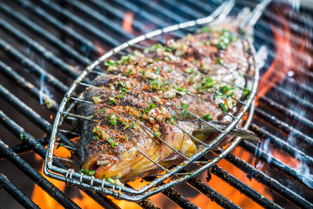 Whole fish on a grill with seasoning and herbs, cooking over an open flame.
