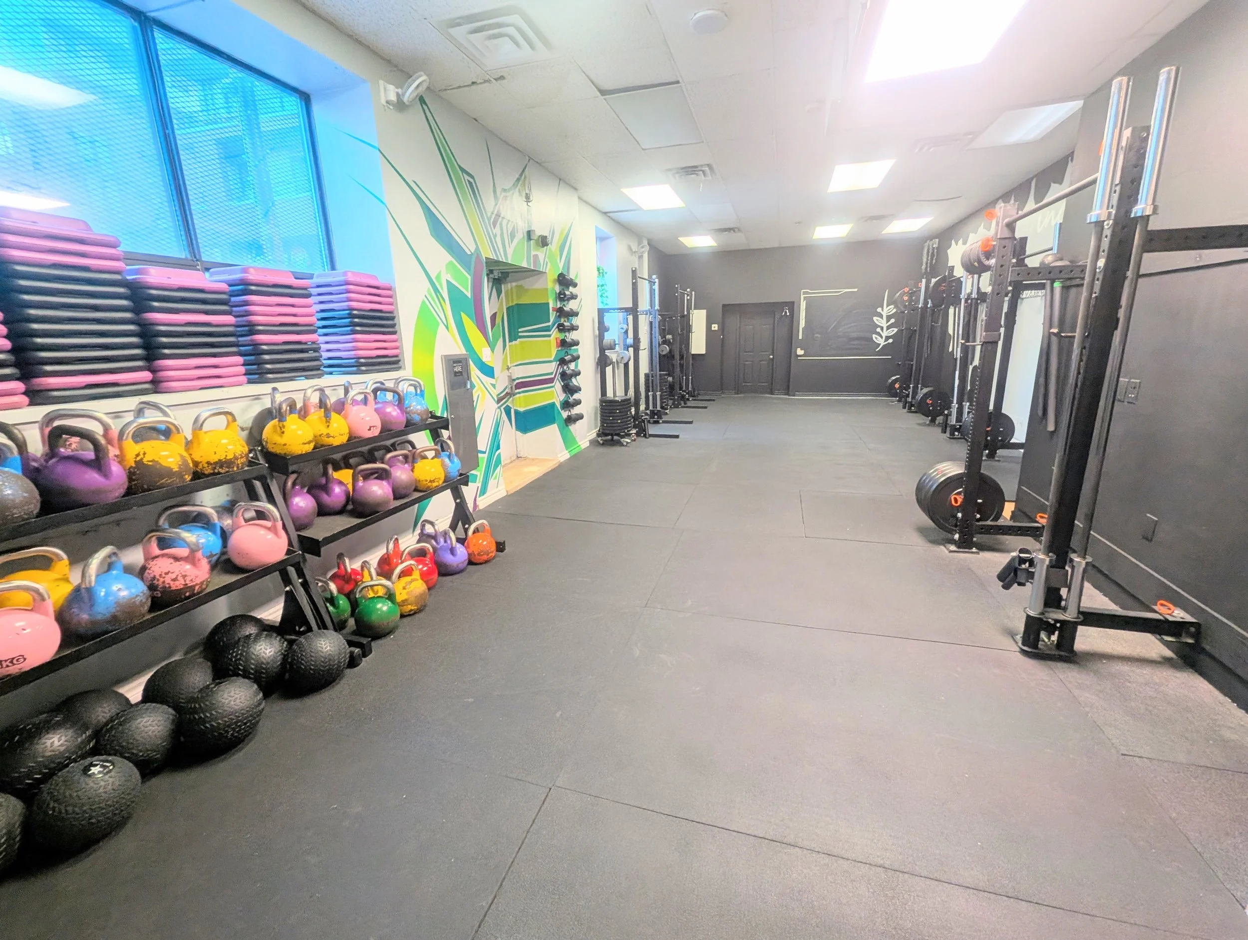 A gym with colorful kettlebells on racks, stacked weight plates, and various weightlifting equipment against the walls. The room has black flooring, gray walls, and windows with blue tint.