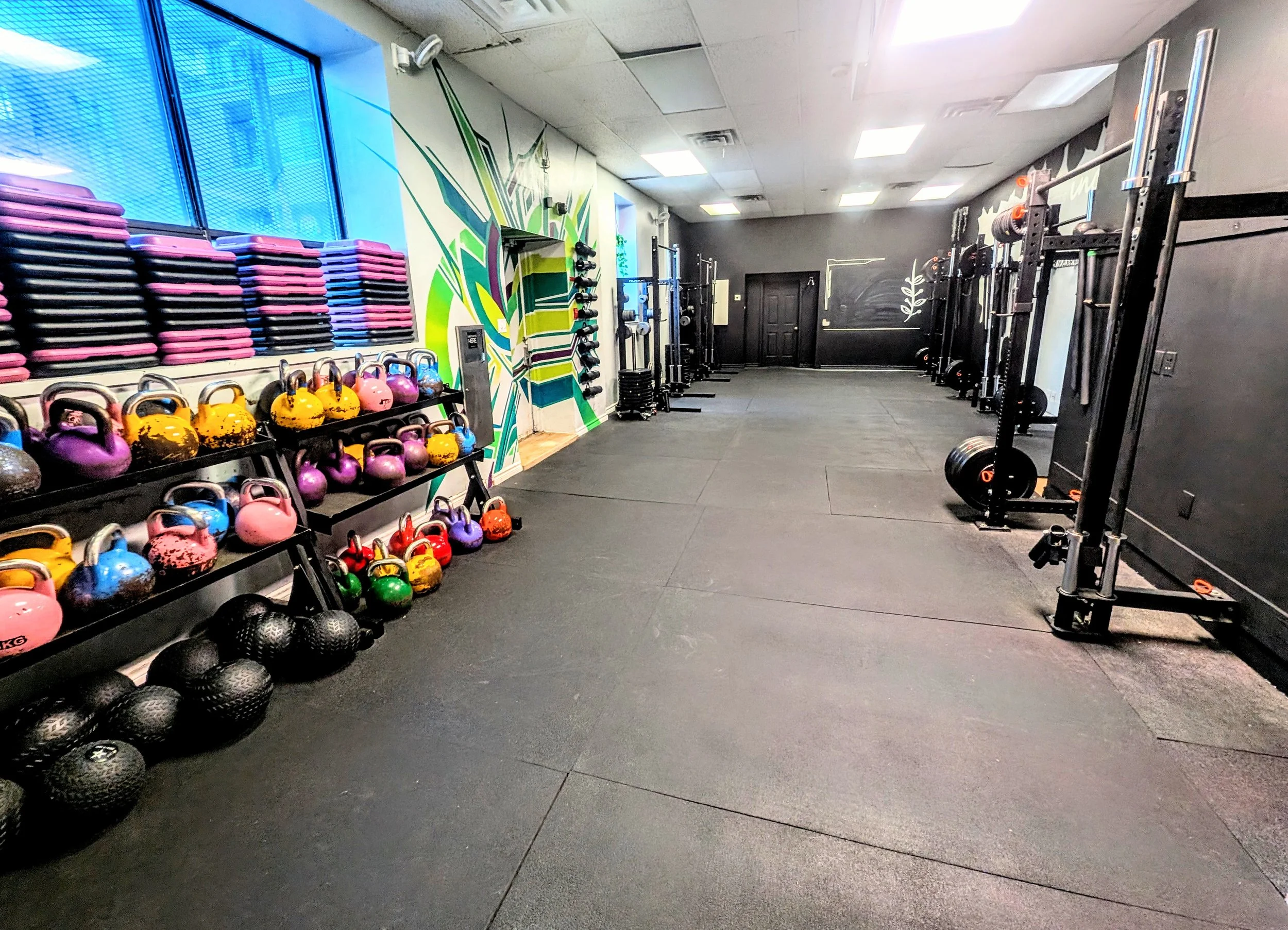 Empty gym with black rubber flooring, colorful kettlebells, medicine balls, stacked pink and purple weight plates, and black weightlifting equipment along the walls. Bright overhead lights and wall art are visible.