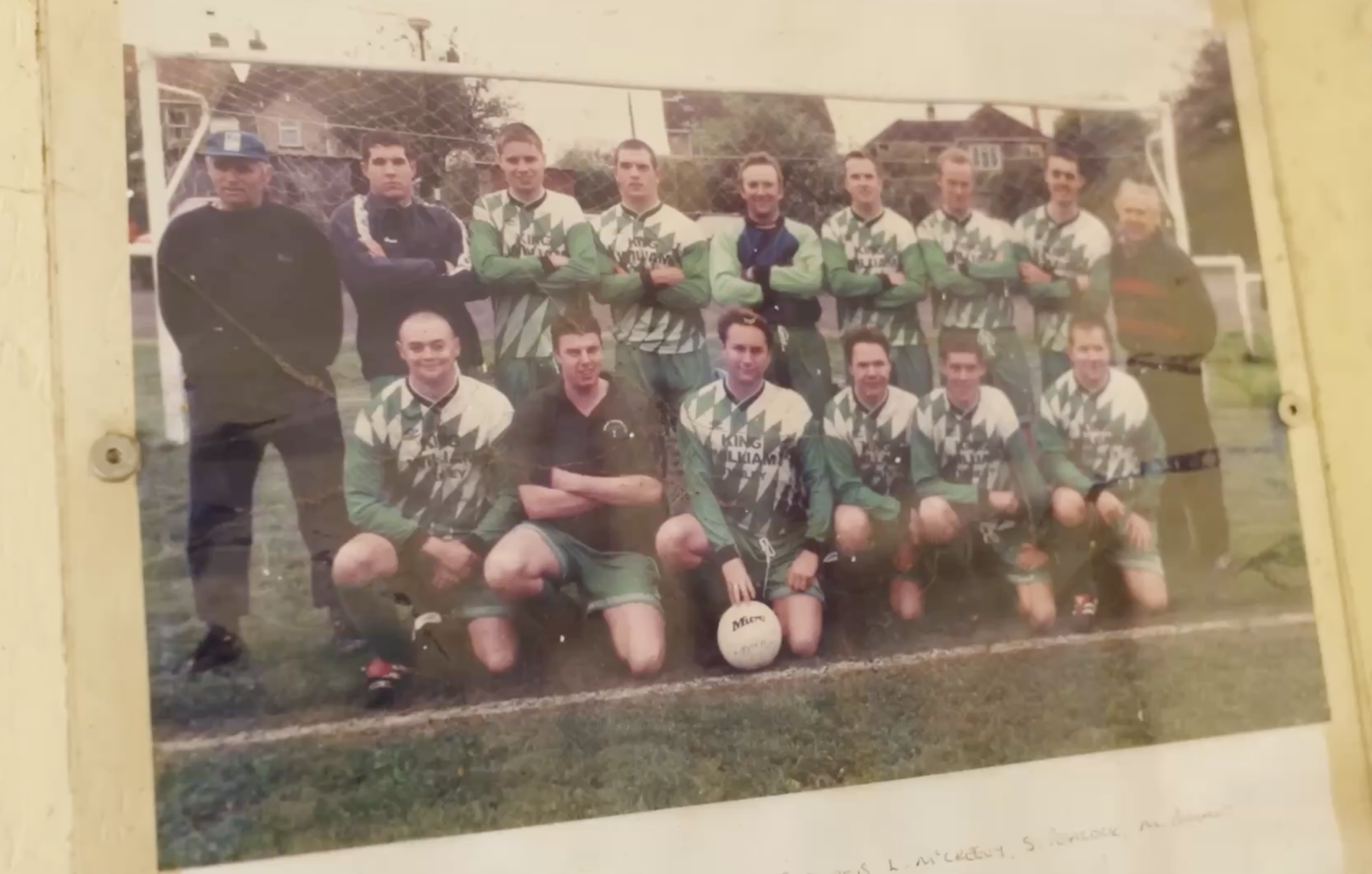 A vintage soccer team photo showing 14 players and 2 coaches or staff members, on a grassy field with a goalpost in the background. The team is in green and black jerseys with a lightning bolt pattern, and the players are kneeling and standing in two