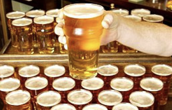 A person holding a pint of beer with a frothy head in front of rows of unopened beer bottles or cans on a bar or countertop.