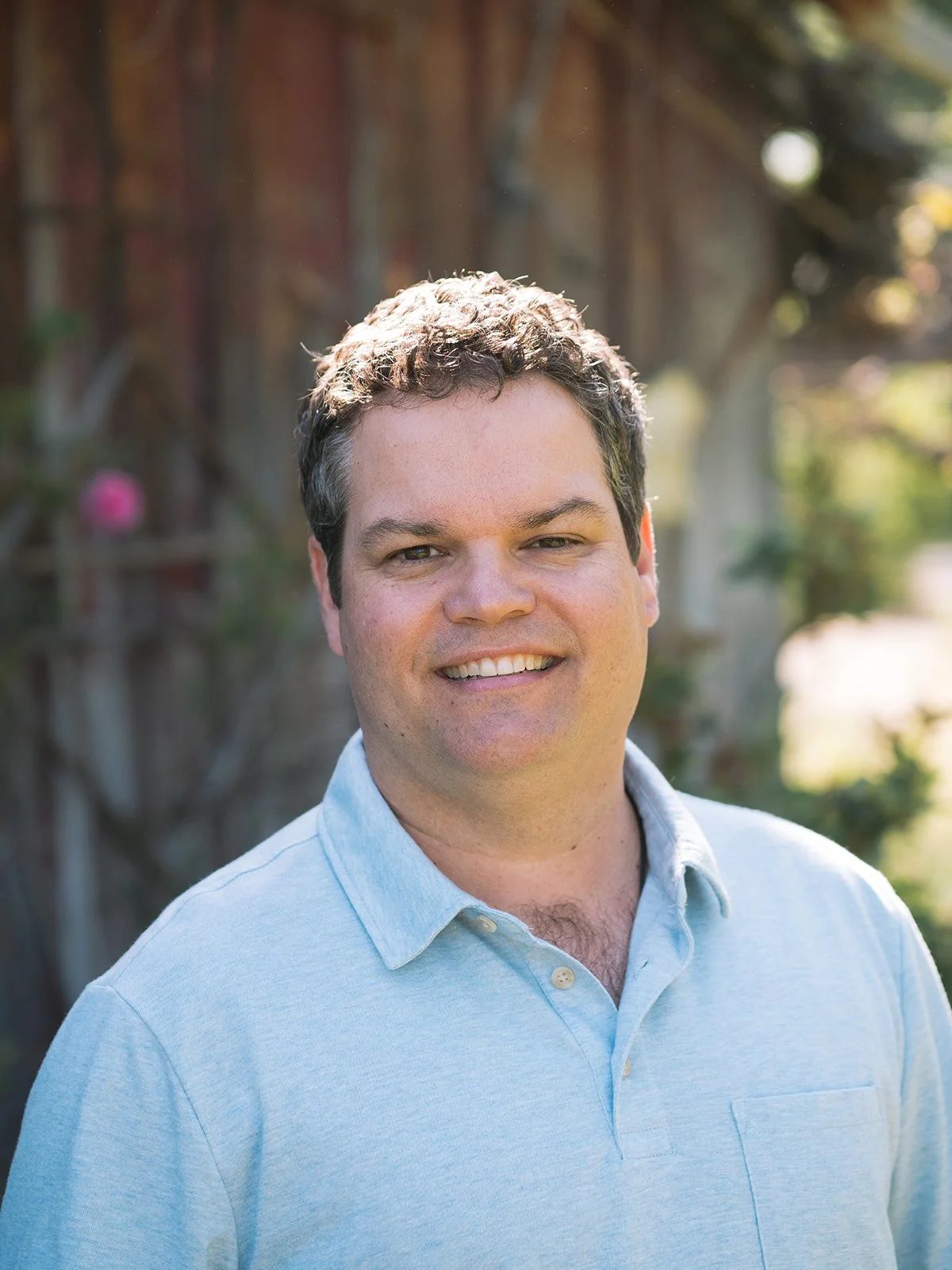 A smiling man with dark, curly hair wearing a light blue collared shirt outdoors with sunlight and blurred greenery in the background.