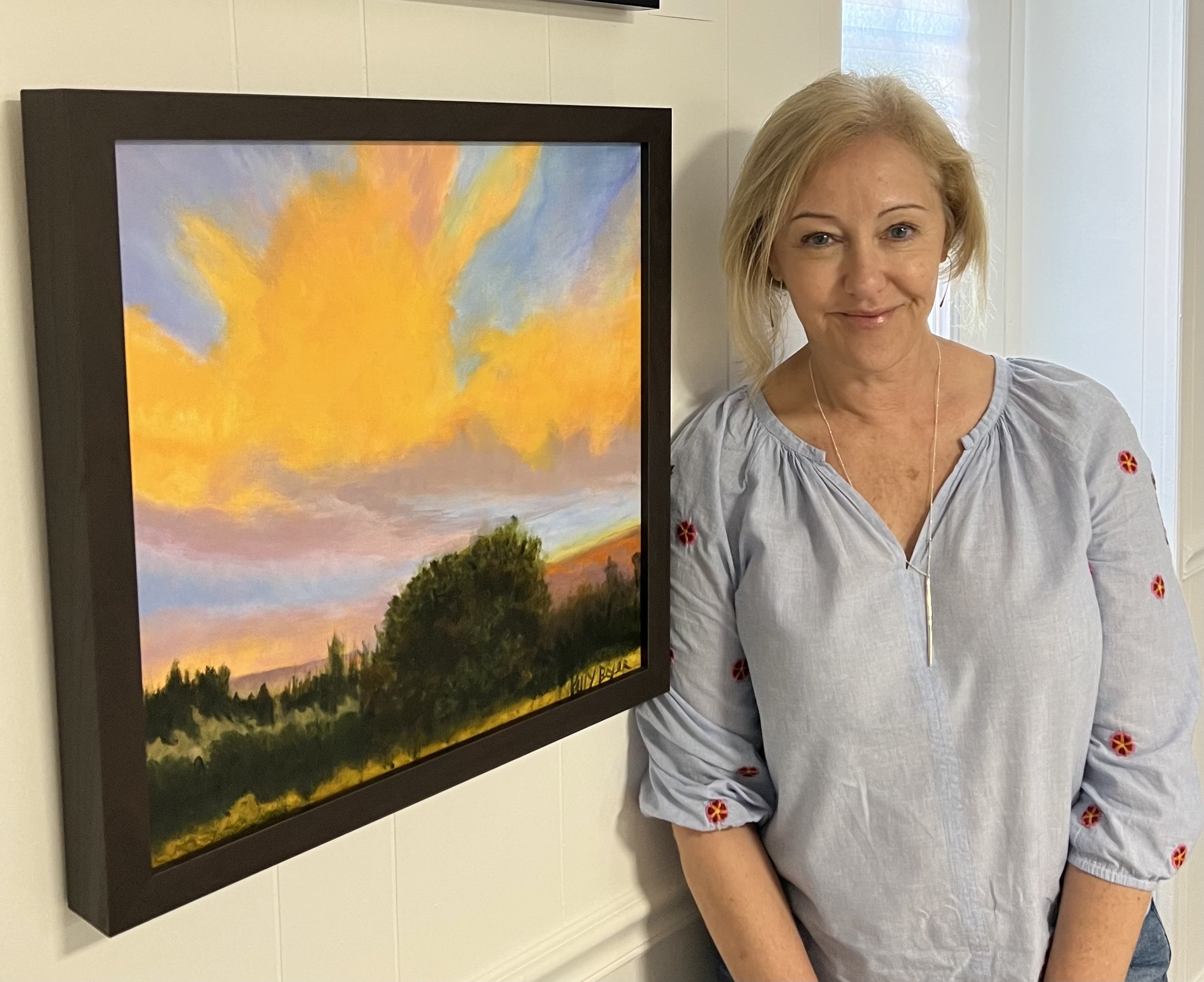 A woman standing next to a framed painting of a colorful sunset landscape, smiling at the camera.