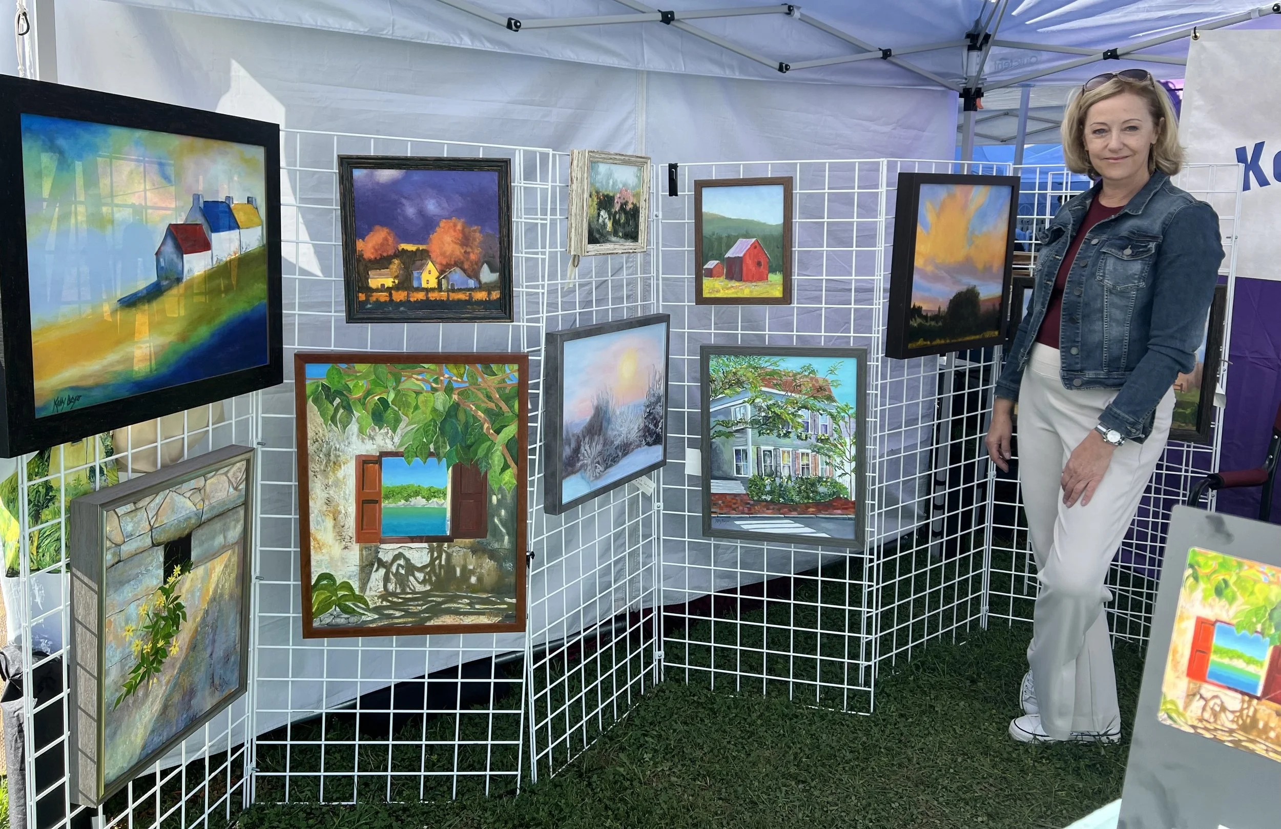 An outdoor art display booth with colorful landscape paintings on a metal grid. A woman in a denim jacket and white pants stands next to the artwork, smiling.
