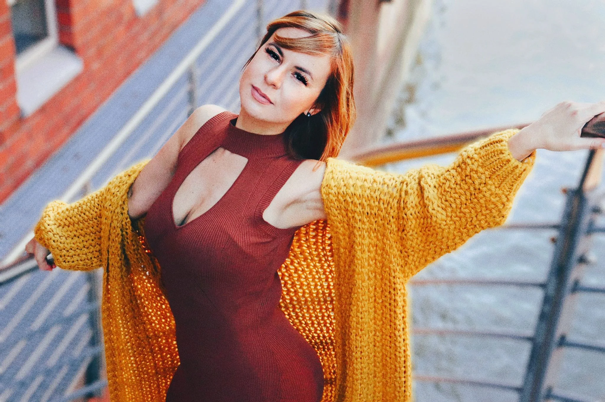 Junge Frau mit braunen Haaren, in rotem Kleid und gelber Strickjacke, steht auf einer Brücke neben Wasser und lächelt in die Kamera.