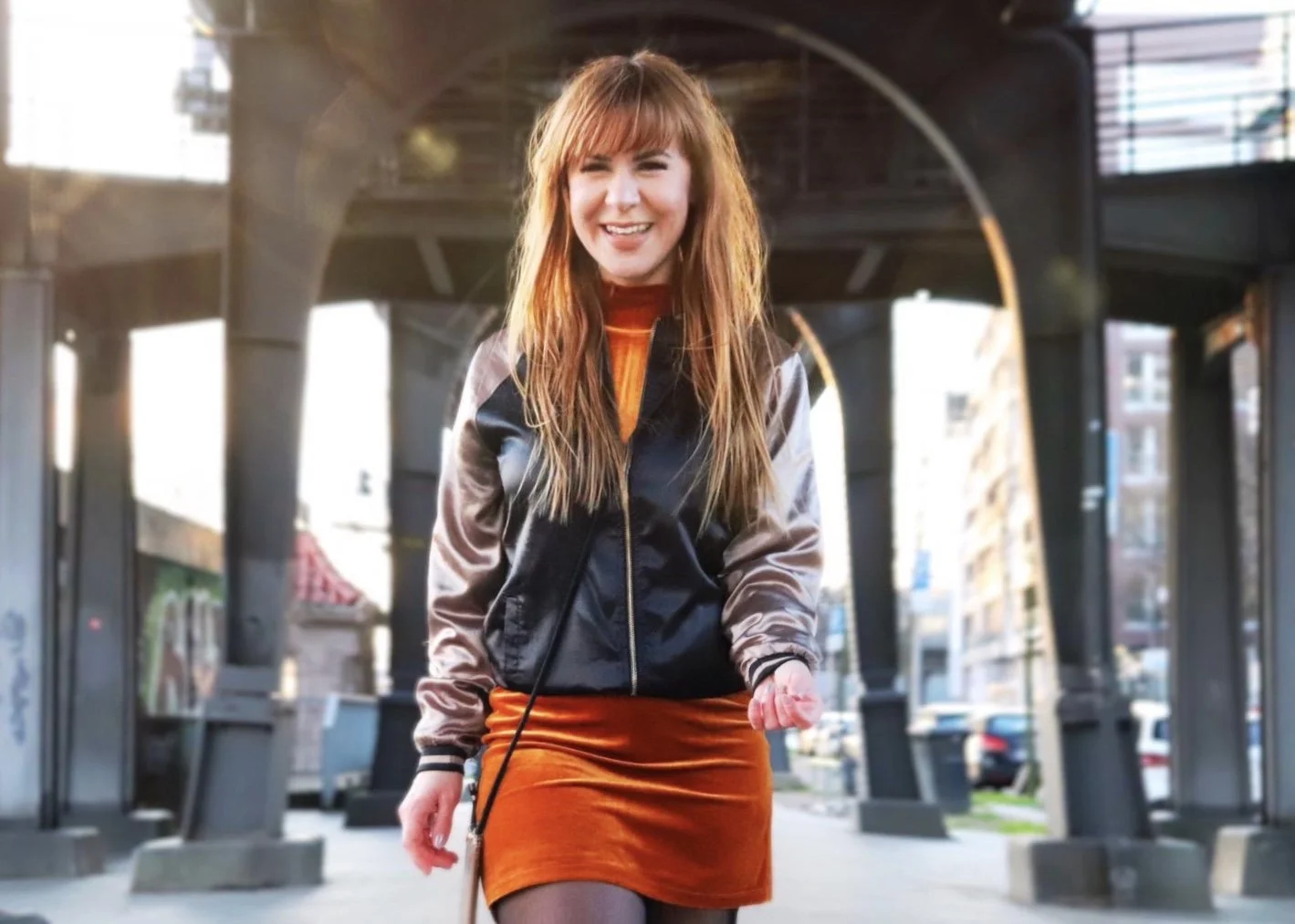 Lächelnde junge Frau in einer Innenstadtunterführung mit modernen Gebäuden im Hintergrund, trägt einen bunten Rock und eine Jacke.