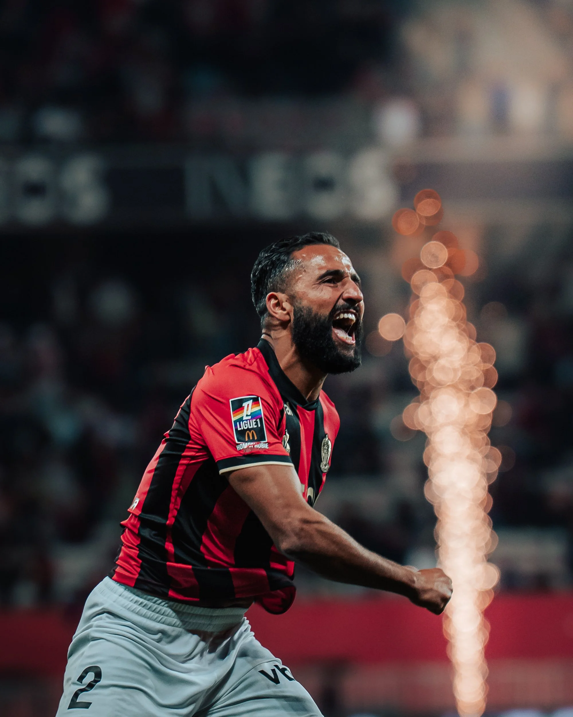 Un homme en maillot de football rouge et noir célèbre sur un terrain, avec des lumières floues en arrière-plan.