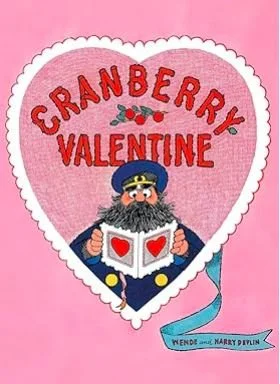 A Valentine card with a pink background and a white scalloped border. It says 'CRANBERRY VALENTINE' in red letters inside a heart-shaped design. There is a cartoon character resembling a pirate or military officer with a gray beard, wearing a blue hat and navy uniform, holding an open book with a red heart on each page. A blue ribbon beneath has the text 'WENDY AND MARRY DEVLIN.'