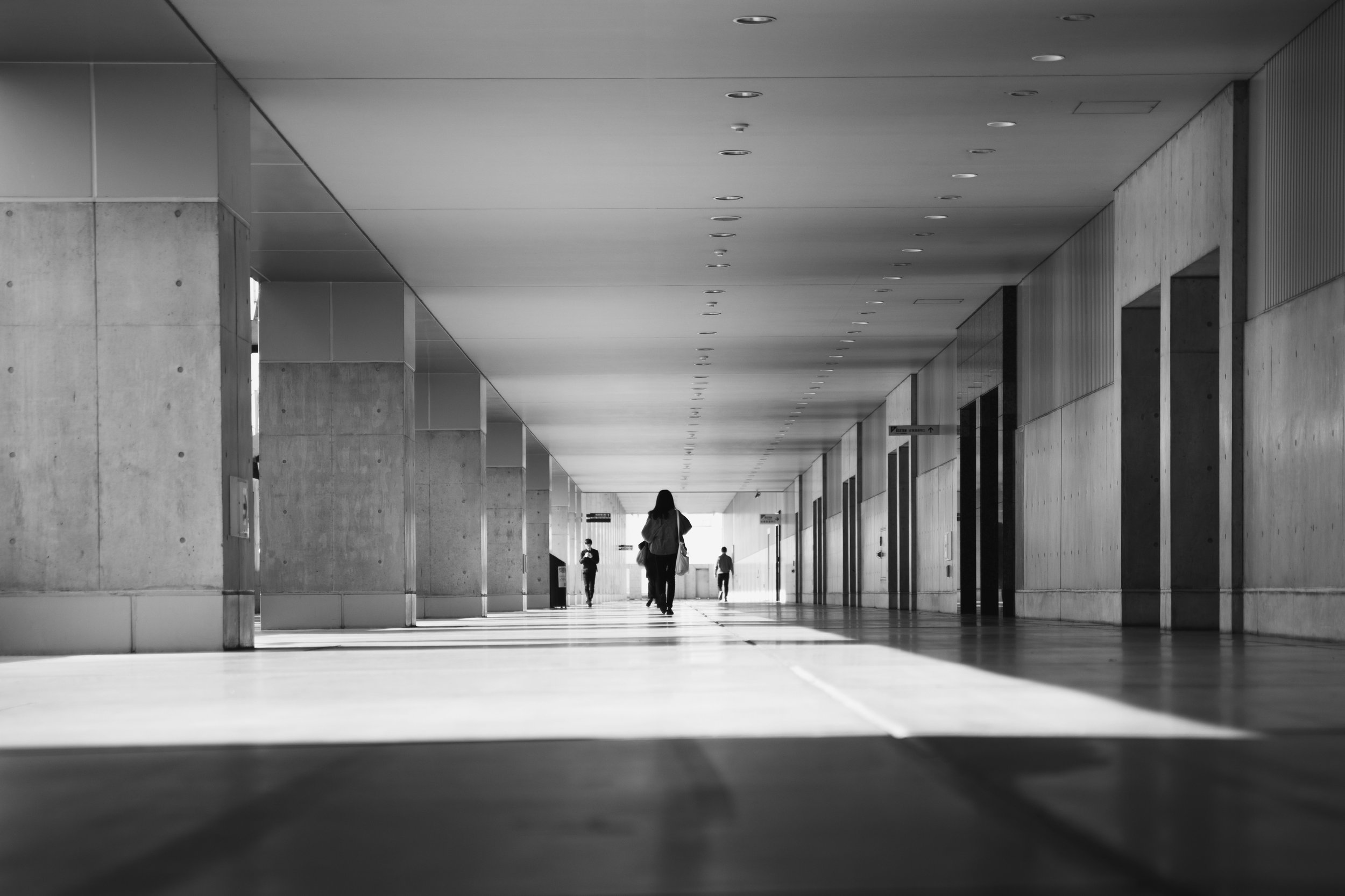 Indoor corridor with concrete walls and ceiling, featuring multiple evenly spaced ceiling lights. Several people are walking away from the camera towards a bright exit at the end of the corridor, with sunlight causing shadows on the floor.