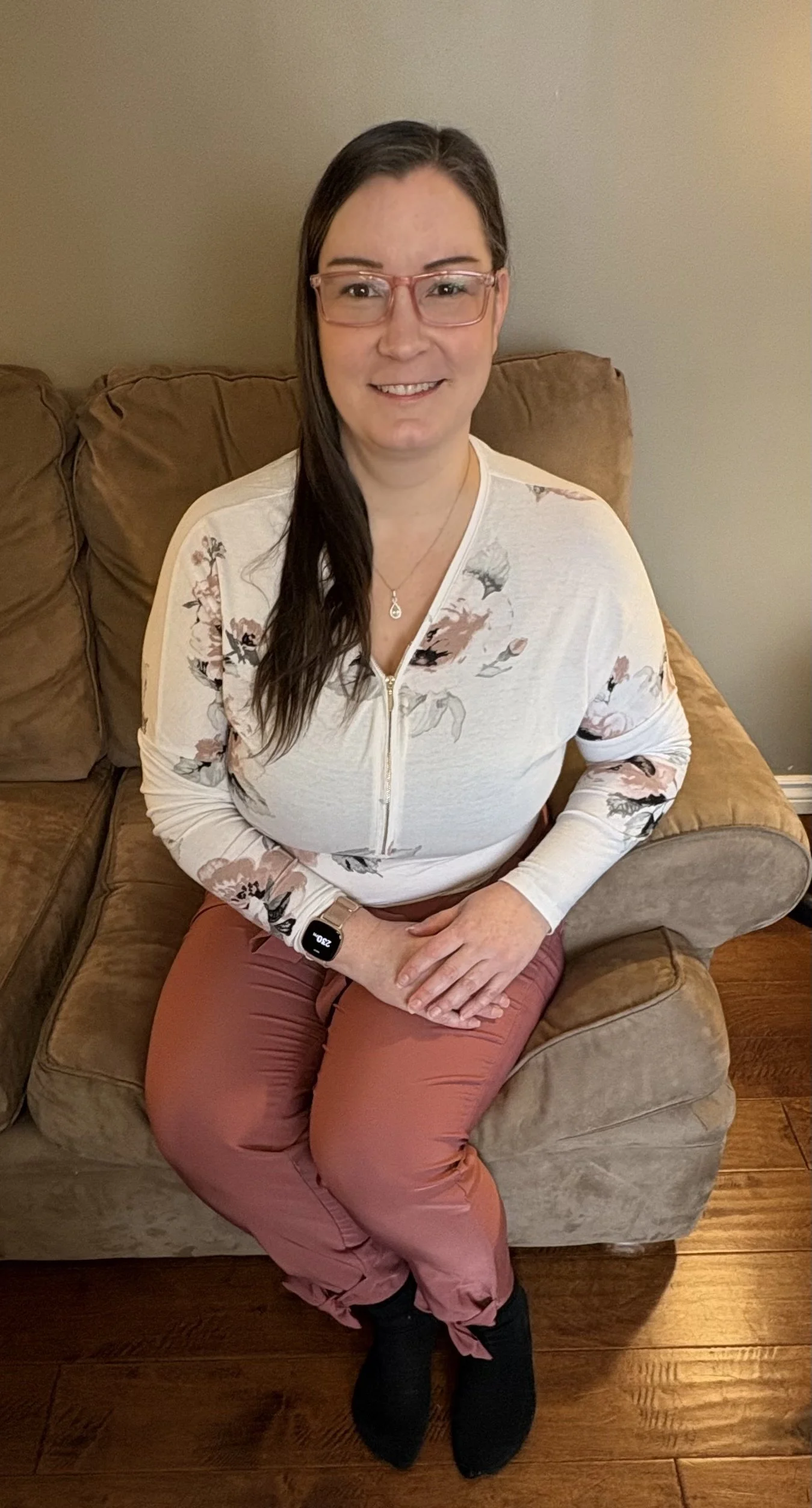 A woman with glasses, long dark hair, wearing a pink top, black cardigan, and patterned pants, sitting on a dark armchair with a white wall in the background, smiling and resting her chin on her hand.