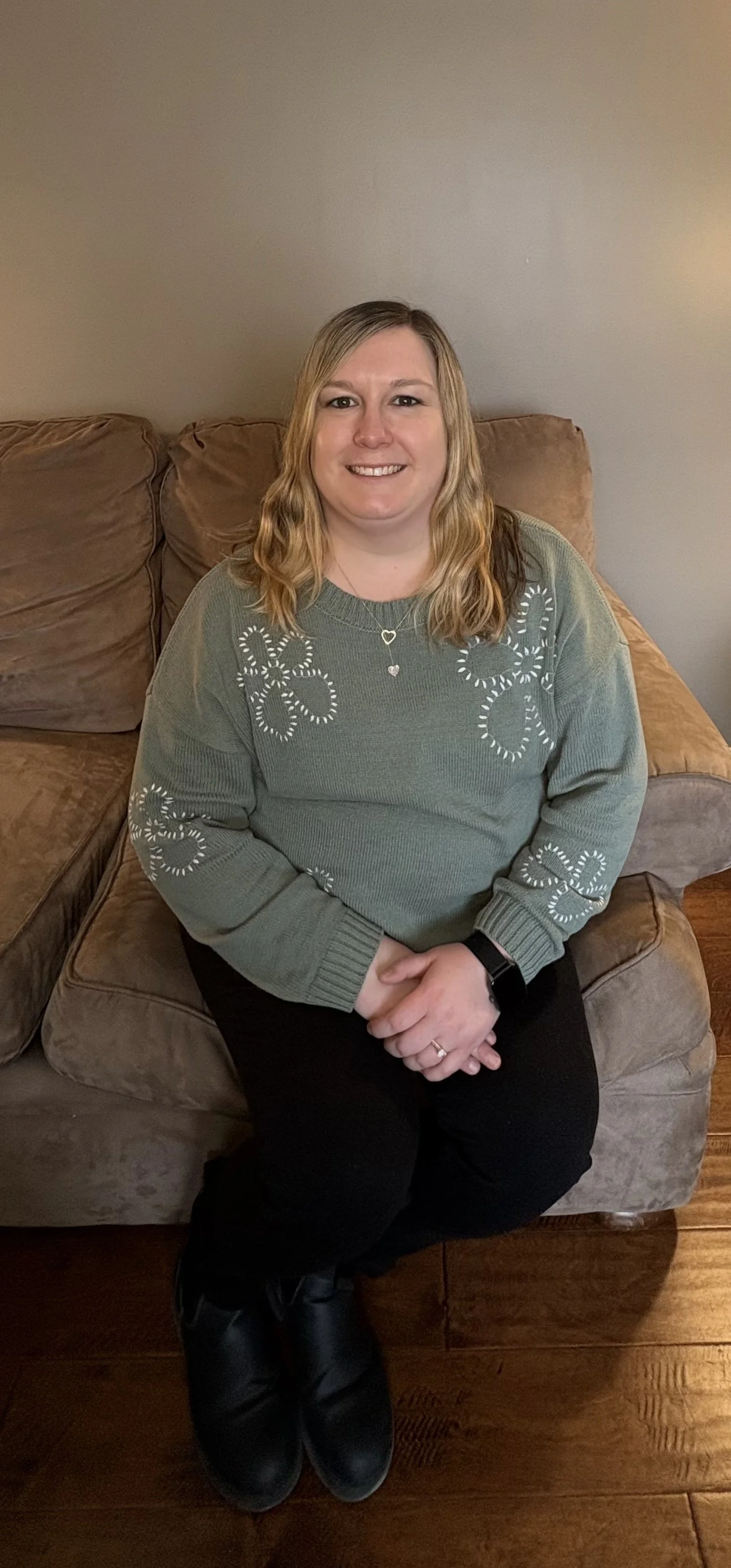 A smiling woman with long blonde hair, wearing a white t-shirt, standing outdoors with a blurred green landscape in the background.