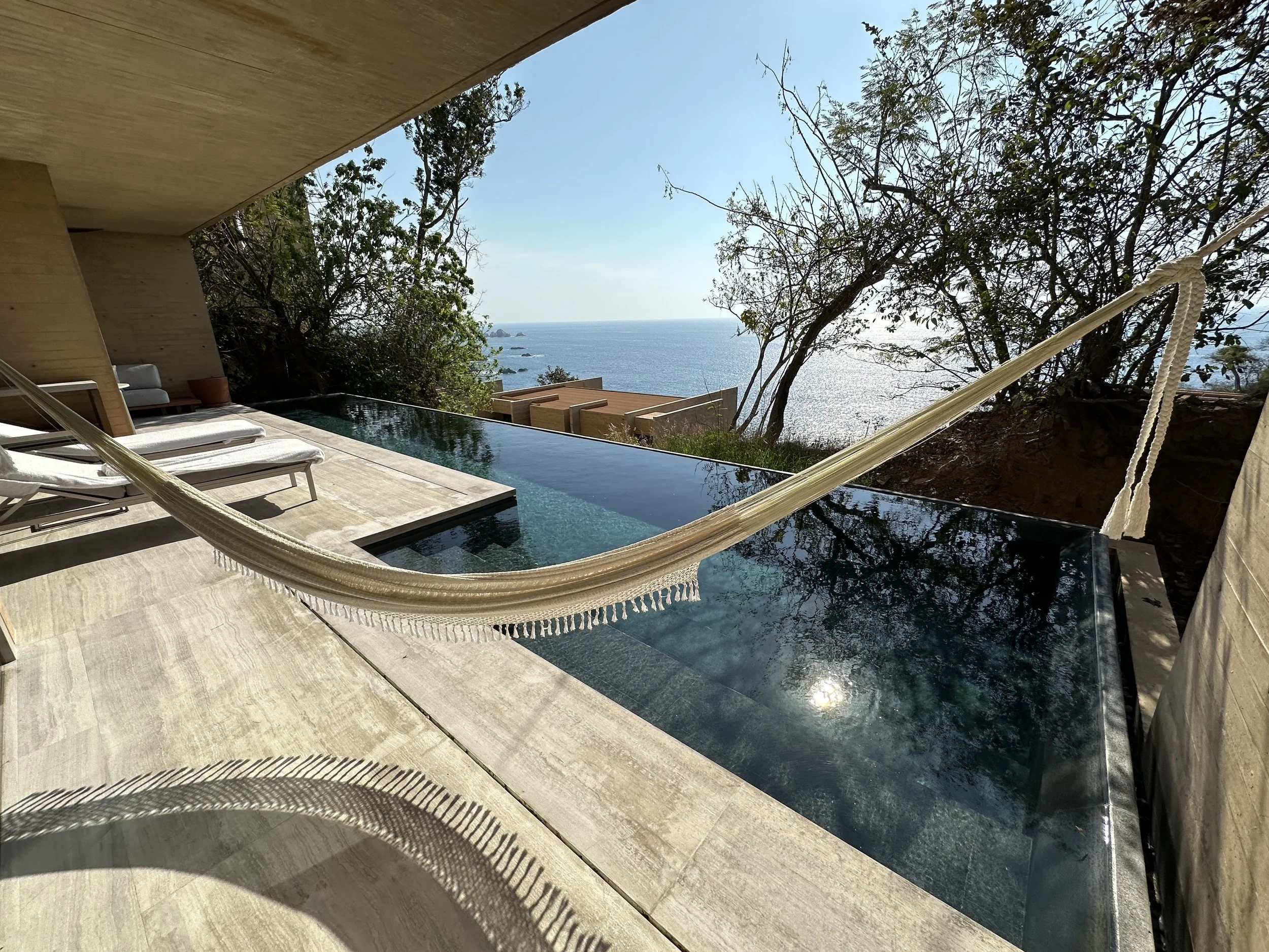 Laura's suite and view at Four Sesons Resort Tamarindo. A modern infinity pool with a view of the ocean, surrounded by lounge chairs and trees, with a hammock hanging nearby.