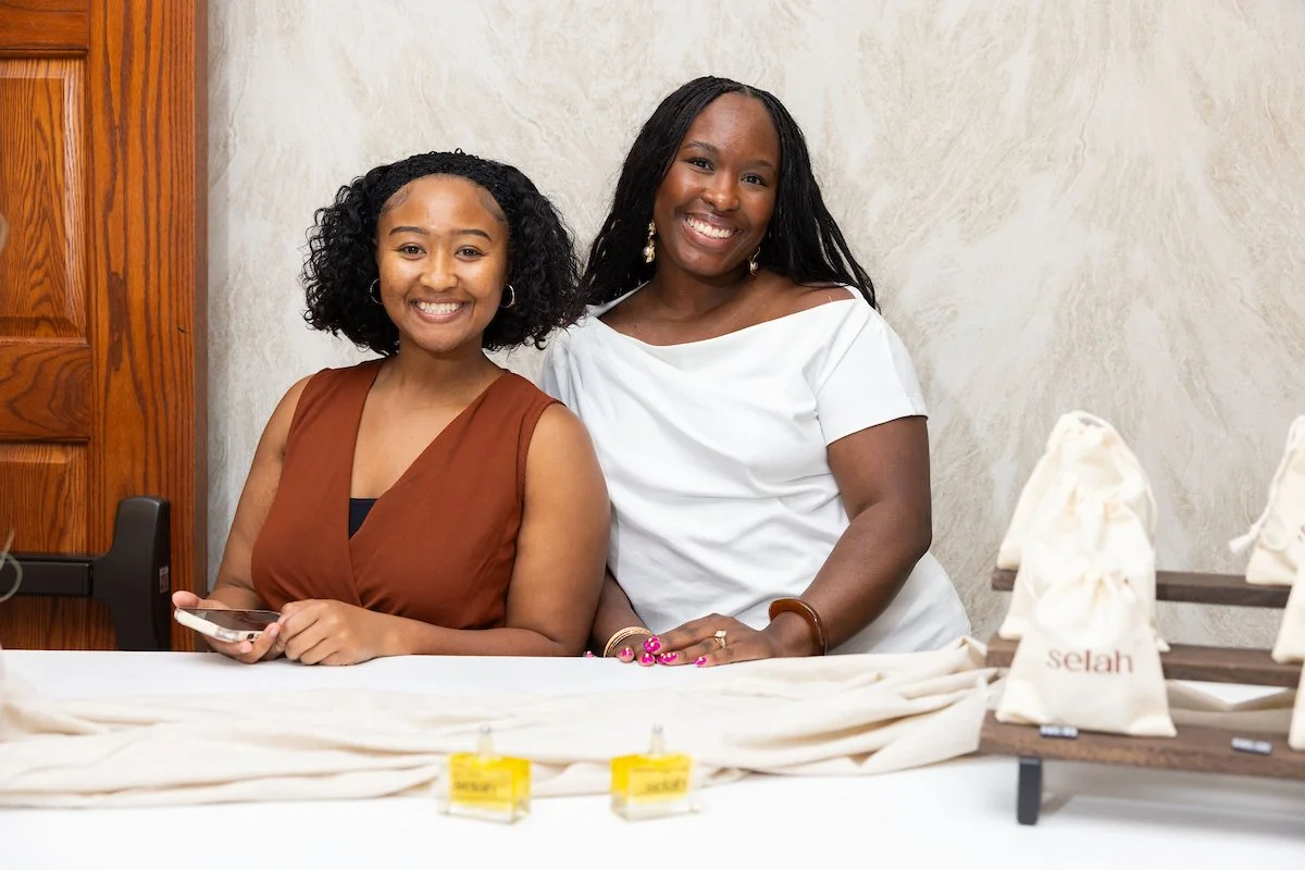 Two women standing behind a check-in table, smiling at the camera. Small items and bags labeled “selah” are displayed on the table in front of them.