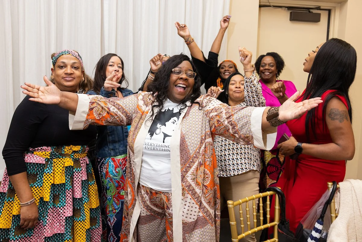 A group of women gathered together, smiling and cheering with arms raised. One woman stands in front with her arms open wide, wearing a patterned outfit and a T-shirt that reads “Everything is Everything.”