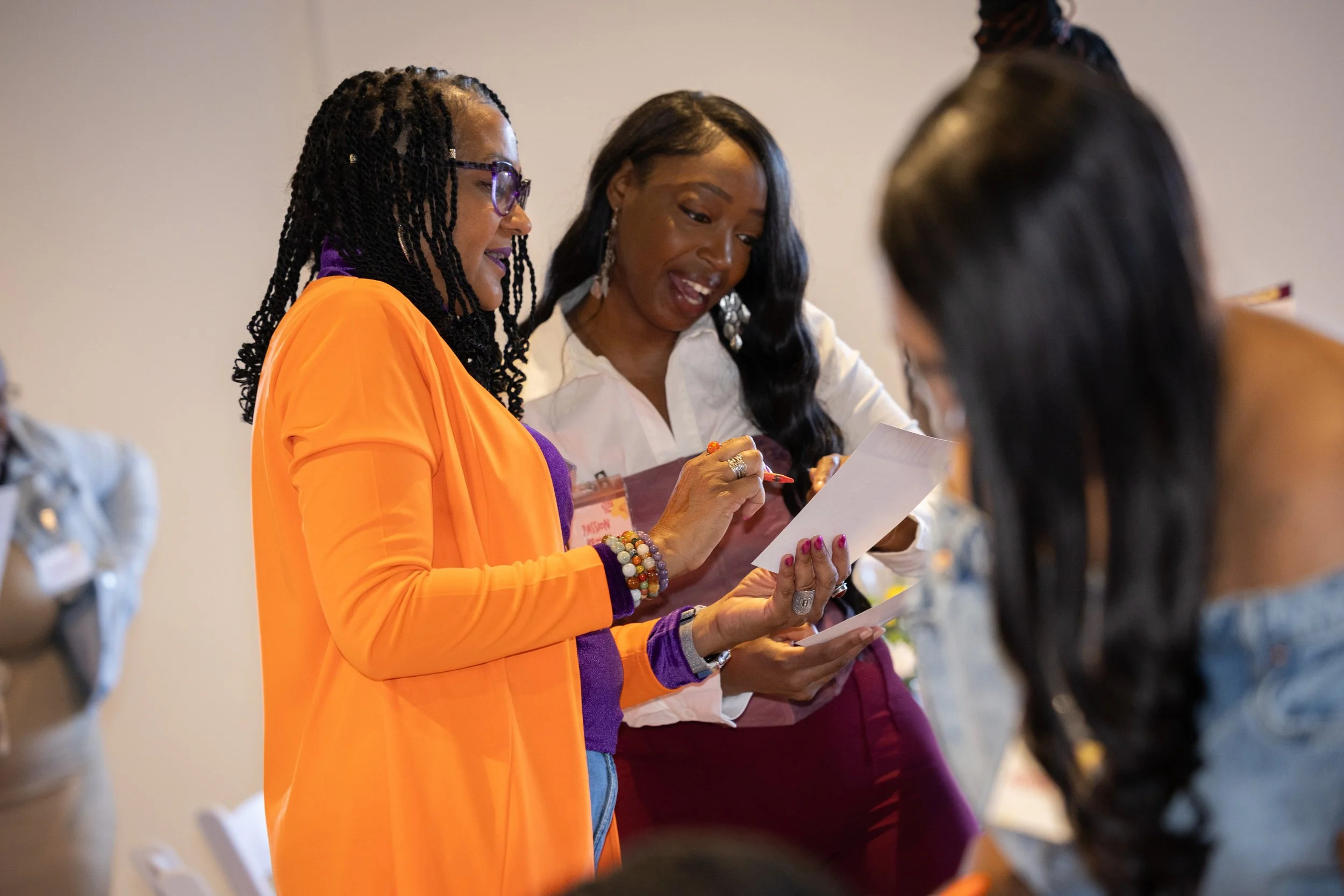 Three women engaged in conversation at a professional or social event, one of them holding a sheet of paper.