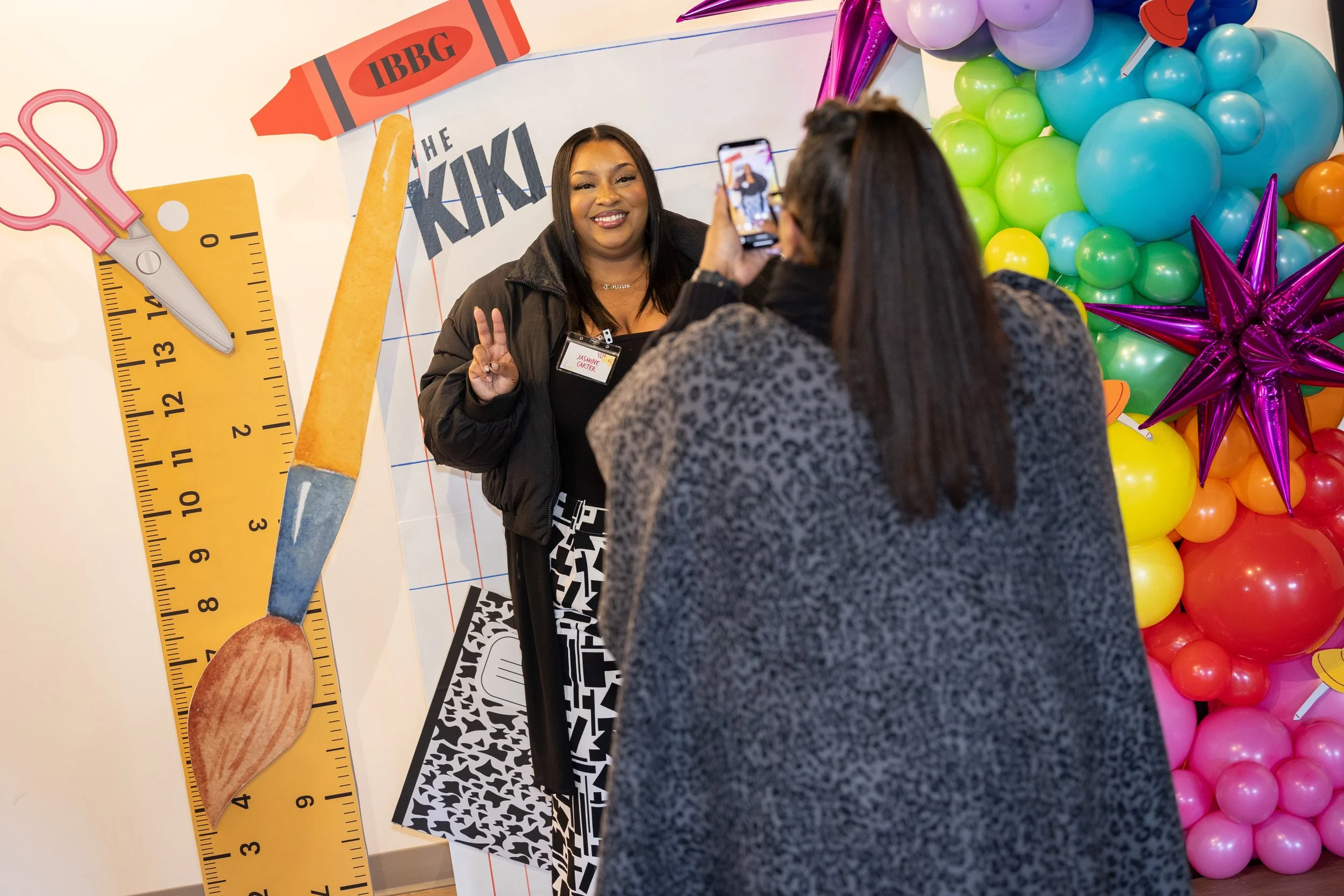 Two women taking a photo of each other at a colorful event with balloon decorations. One woman is smiling and making a peace sign, while the other is holding a phone. The background has a large balloon arrangement and a decorative wall with school su