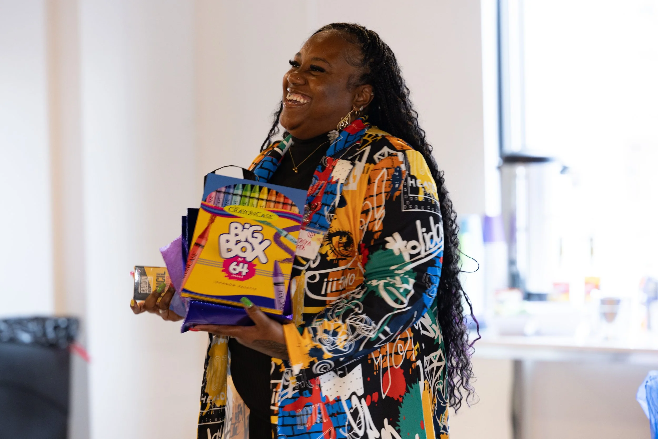 A woman with long, curly black hair smiling and holding a box of Crayola Big Box 64 crayons and other art supplies in a bright room with a window.