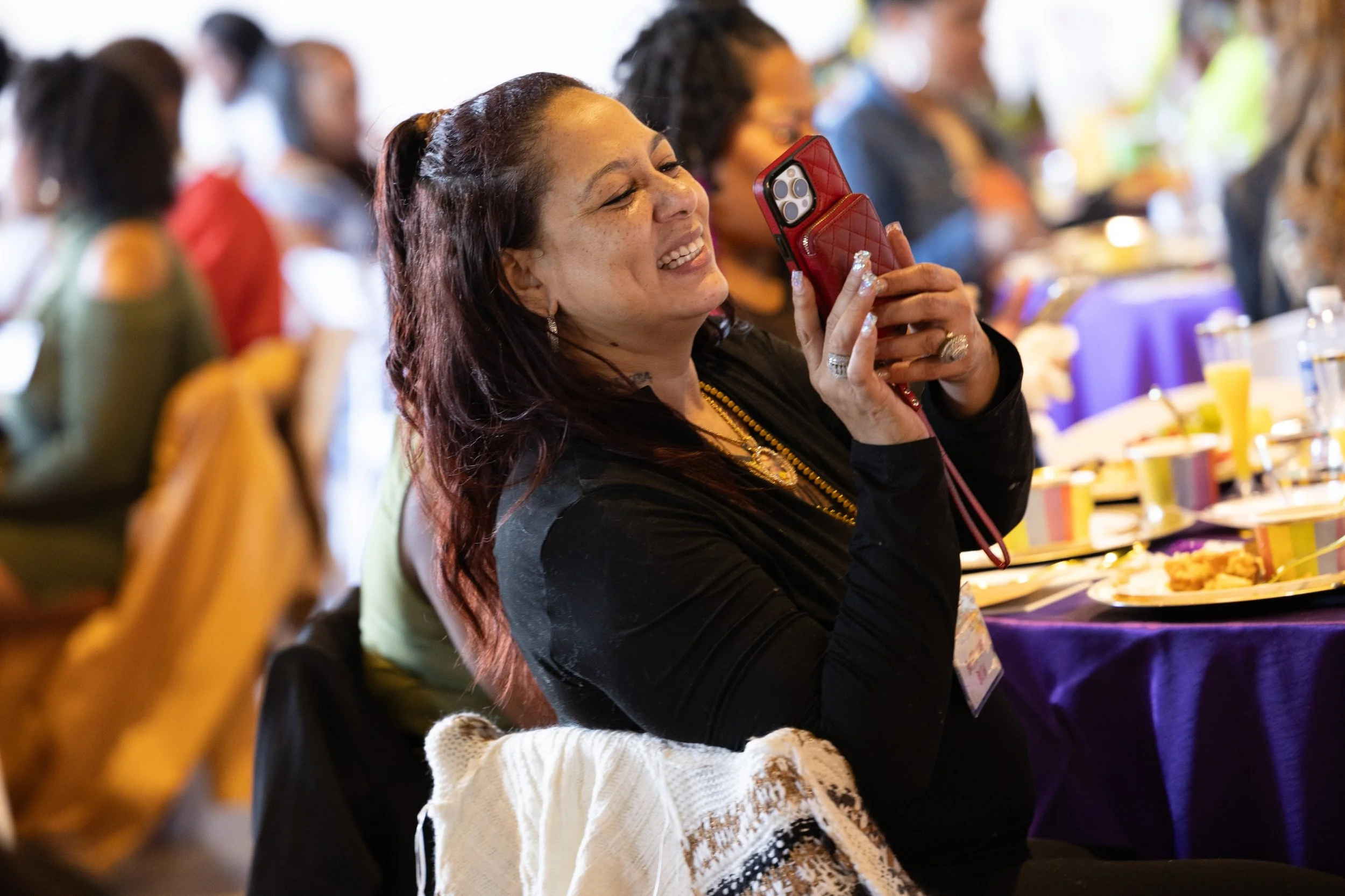 A woman with long dark hair smiling and taking a photo with her phone at a crowded event with many people seated at tables.