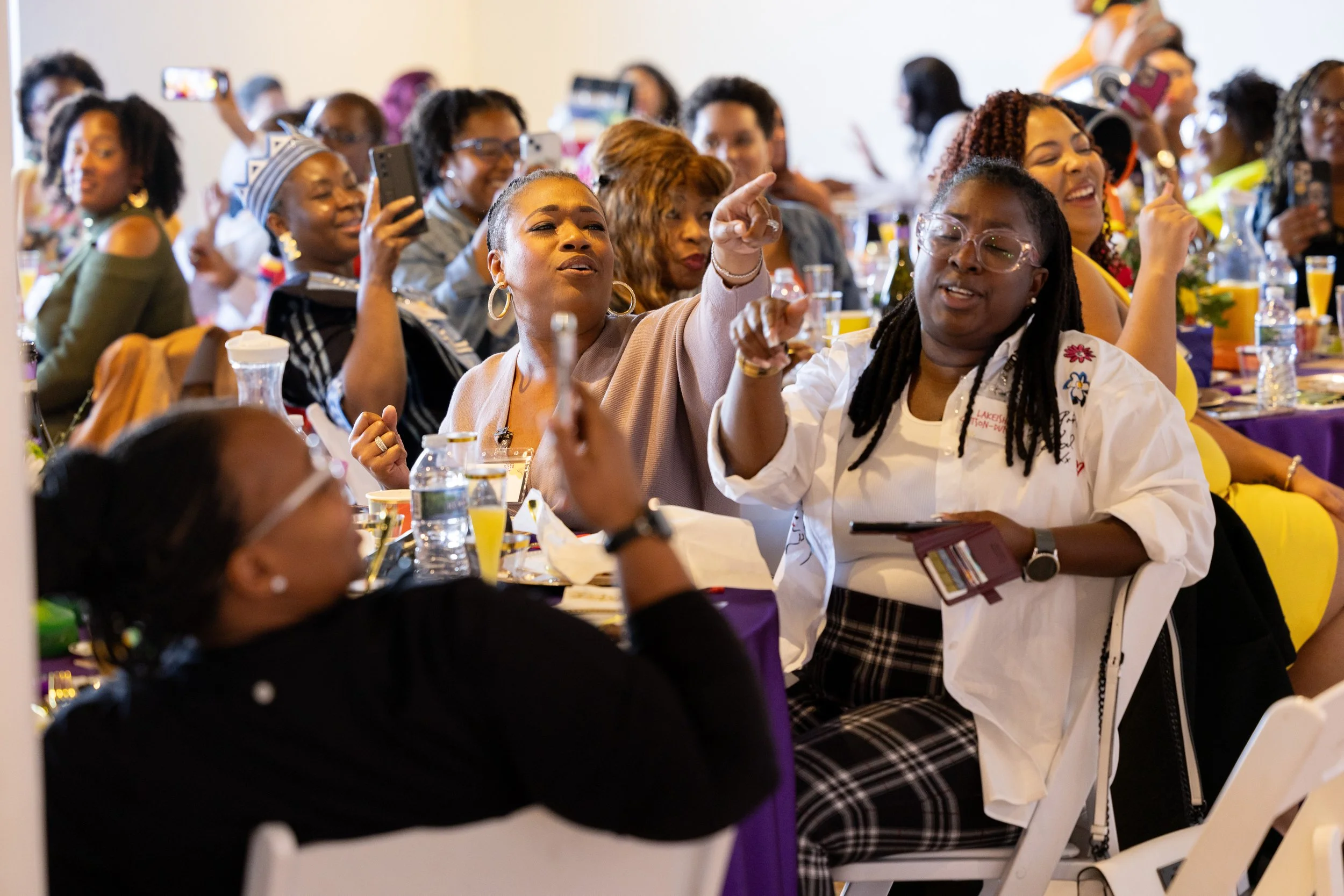 A group of women sitting at a banquet table, engaging and taking photos, at a lively indoor event or conference.