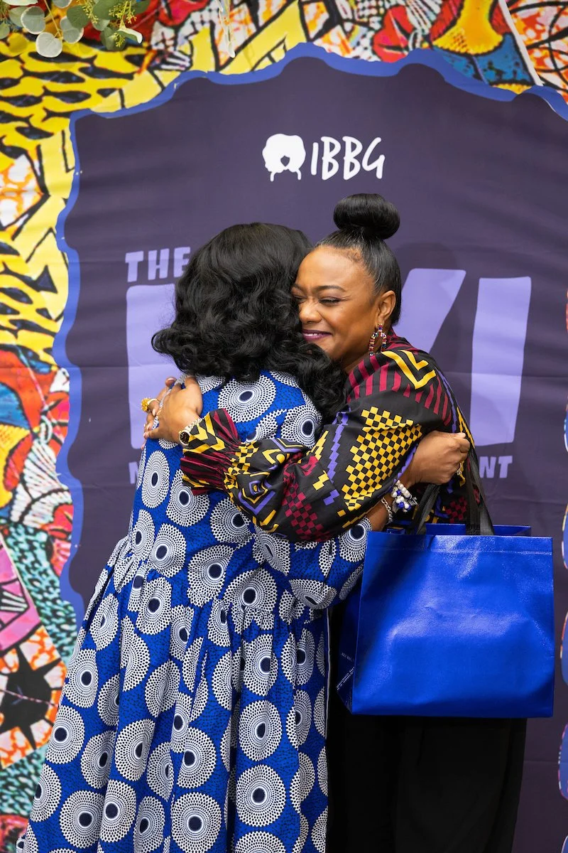 Two women embracing in front of an “IBBG” event backdrop. One holds a large blue bag while they hug warmly.