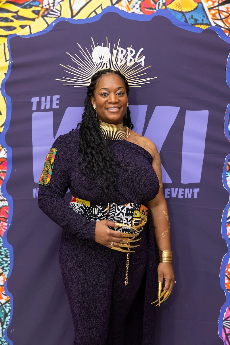 A woman posing in front of an event backdrop with “IBBG” and “The Kiki” visible. She wears a deep purple outfit with gold accessories and stands confidently, smiling.