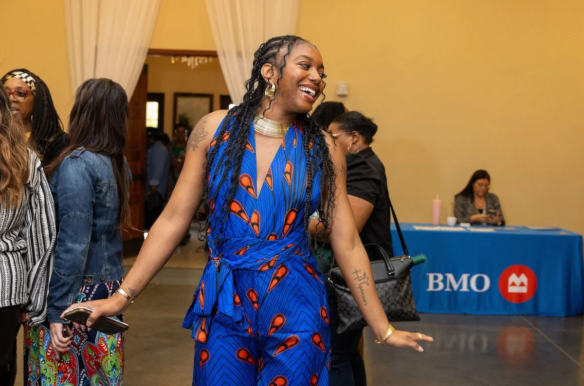 A woman in a blue patterned jumpsuit smiling and moving as if dancing in a crowded event space. Other attendees and a table with a blue “BMO” sign are visible in the background.