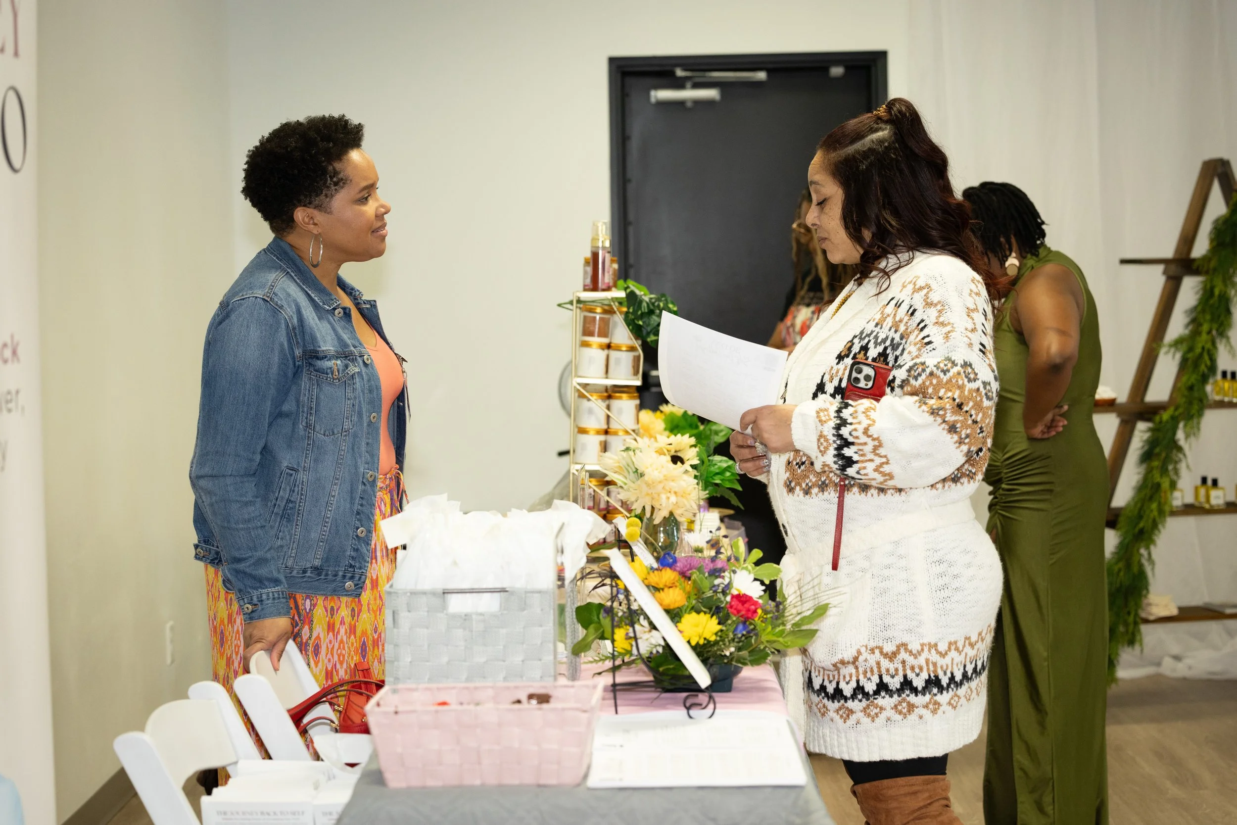 Two women standing at a table with flowers, discussing. The woman on the left has short curly hair and wears a denim jacket, the woman on the right has long dark hair and wears a white patterned sweater. In the background, another woman in a green dr