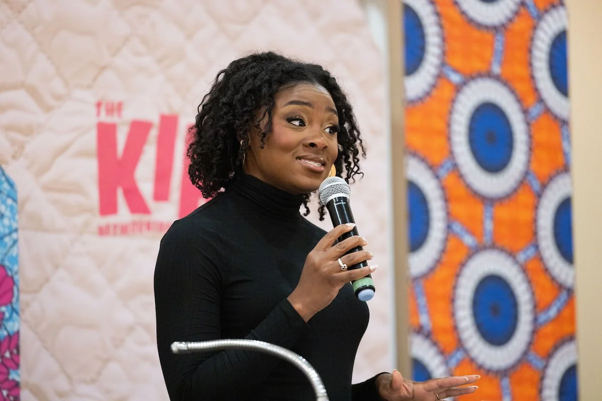 A woman speaking into a microphone at a podium, wearing a black top. Behind her is a colorful patterned backdrop and signage for the event.