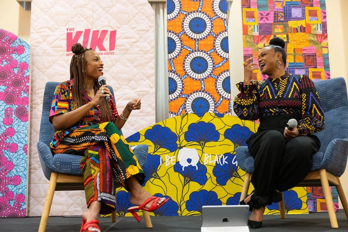 Two women seated on stage in conversation, both holding microphones and smiling. They wear bold, colorful patterned outfits, with a vibrant quilted backdrop and “The Kiki” branding behind them.