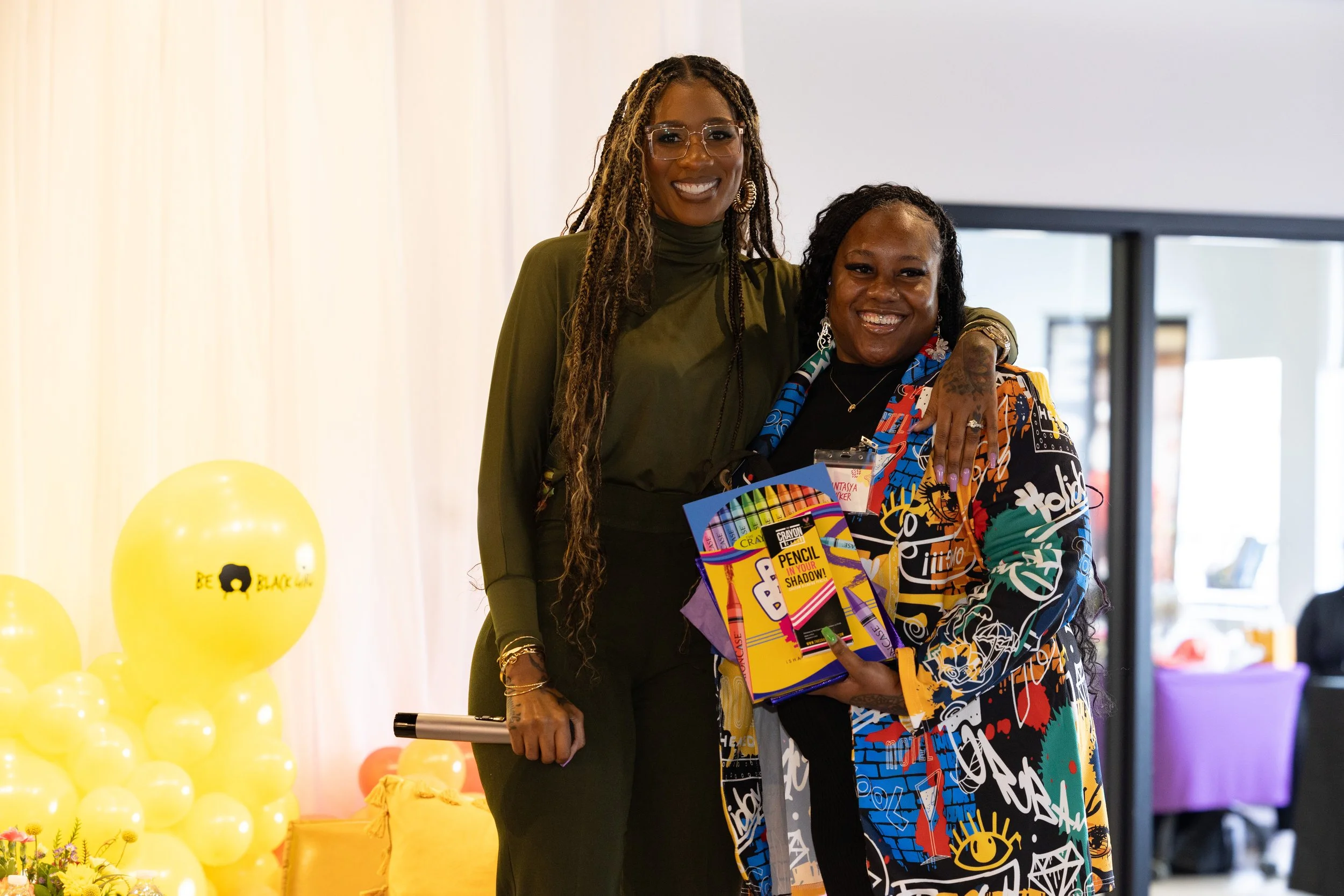 Two women smiling and standing together indoors. One woman is taller with long dreadlocks, wearing glasses and a green dress. The other shorter woman has short hair, wearing a colorful patterned outfit, and holding a pack of Crayola products. There a