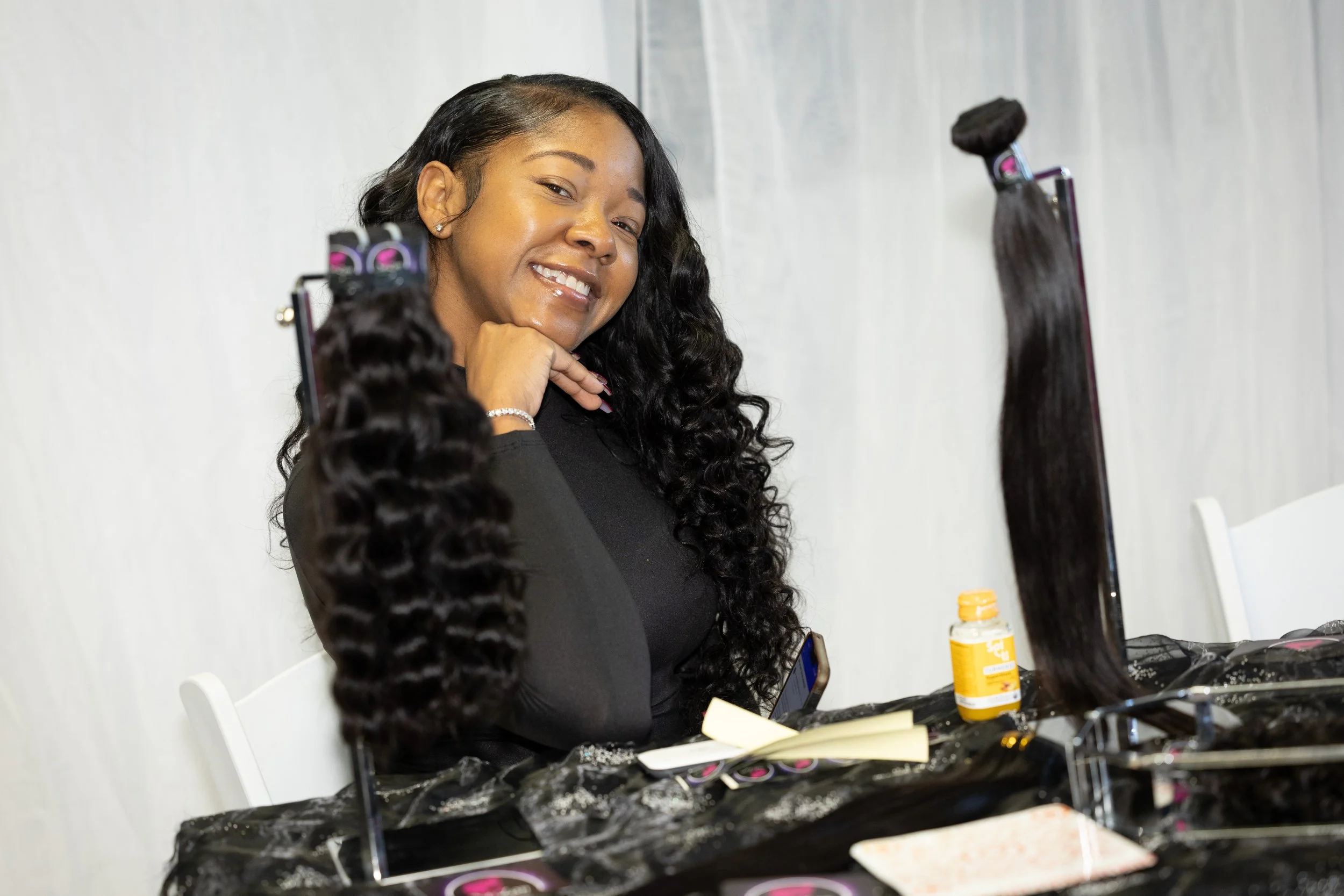 A woman with long wavy black hair, sitting at a table with hair extensions, a notebook, a cell phone, a bottle of vitamins, and a mirror, smiling at the camera.