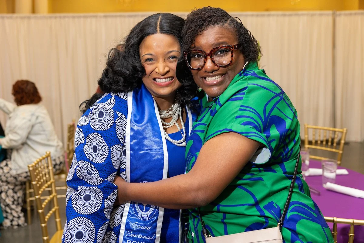Two women smiling and hugging closely for the camera. One wears a blue patterned outfit with a sash, and the other wears a green patterned dress with large glasses. 