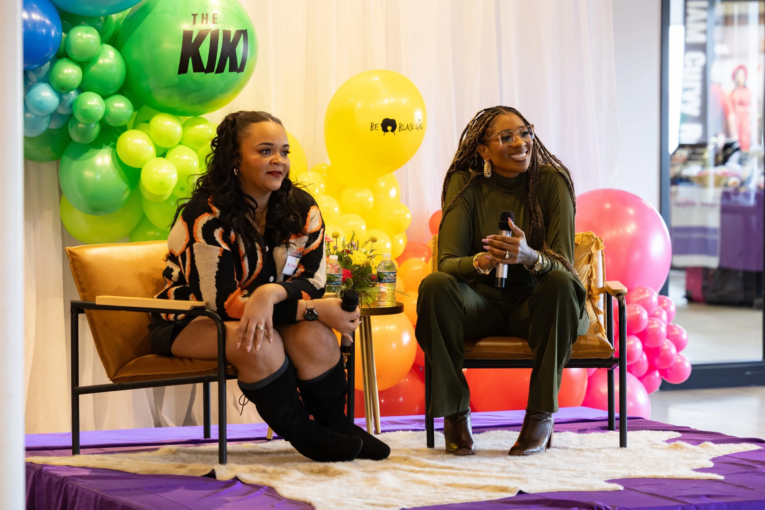 Two women sitting on a stage with balloons behind them. One woman has long dark hair, wearing a patterned sweater, black shorts, and thigh-high boots, holding a microphone. The other woman has long braided hair, glasses, a dark green outfit, and is h