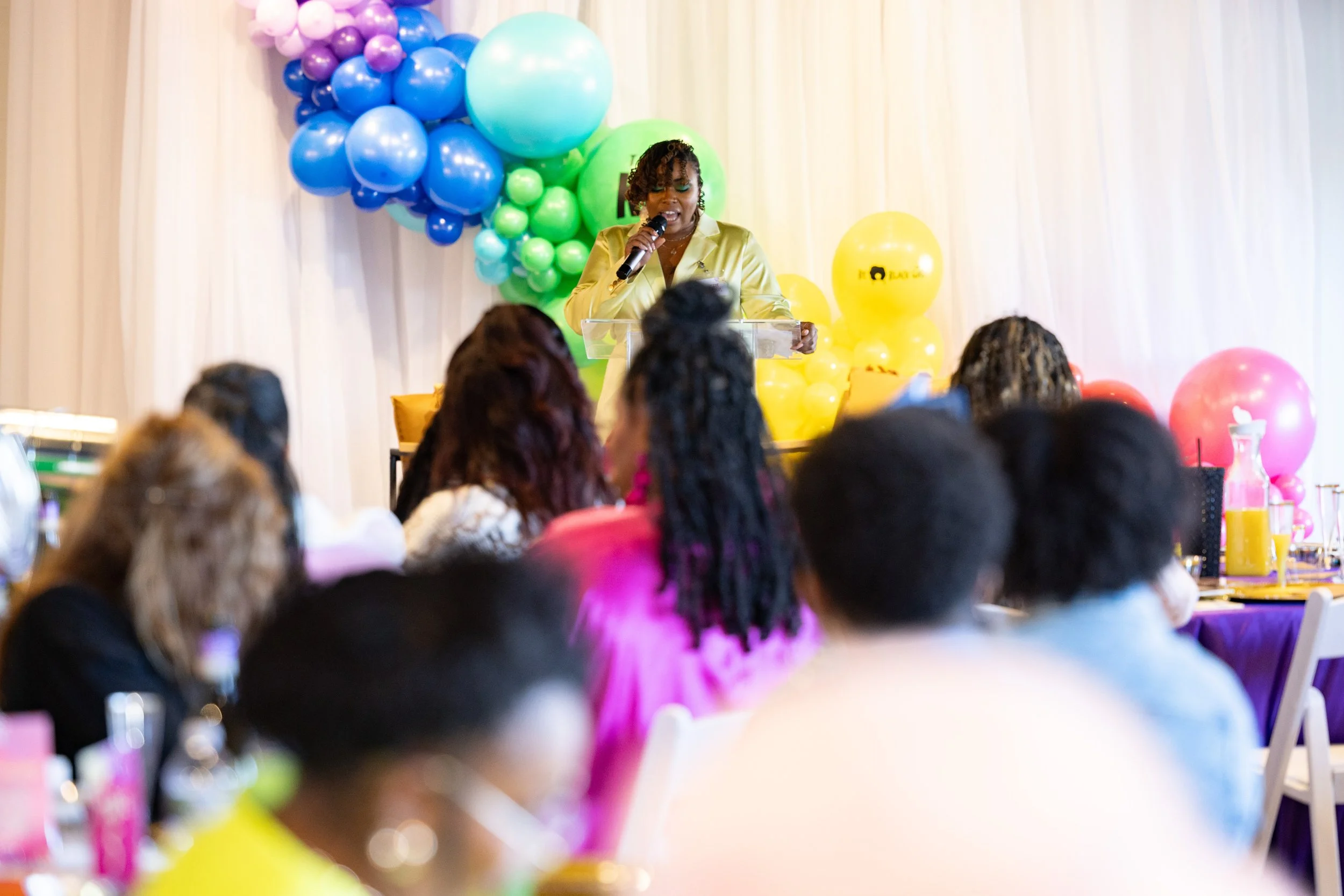 A woman in a yellow jacket speaking at a podium during a gathering with colorful balloons in the background and audience members sitting at decorated tables.