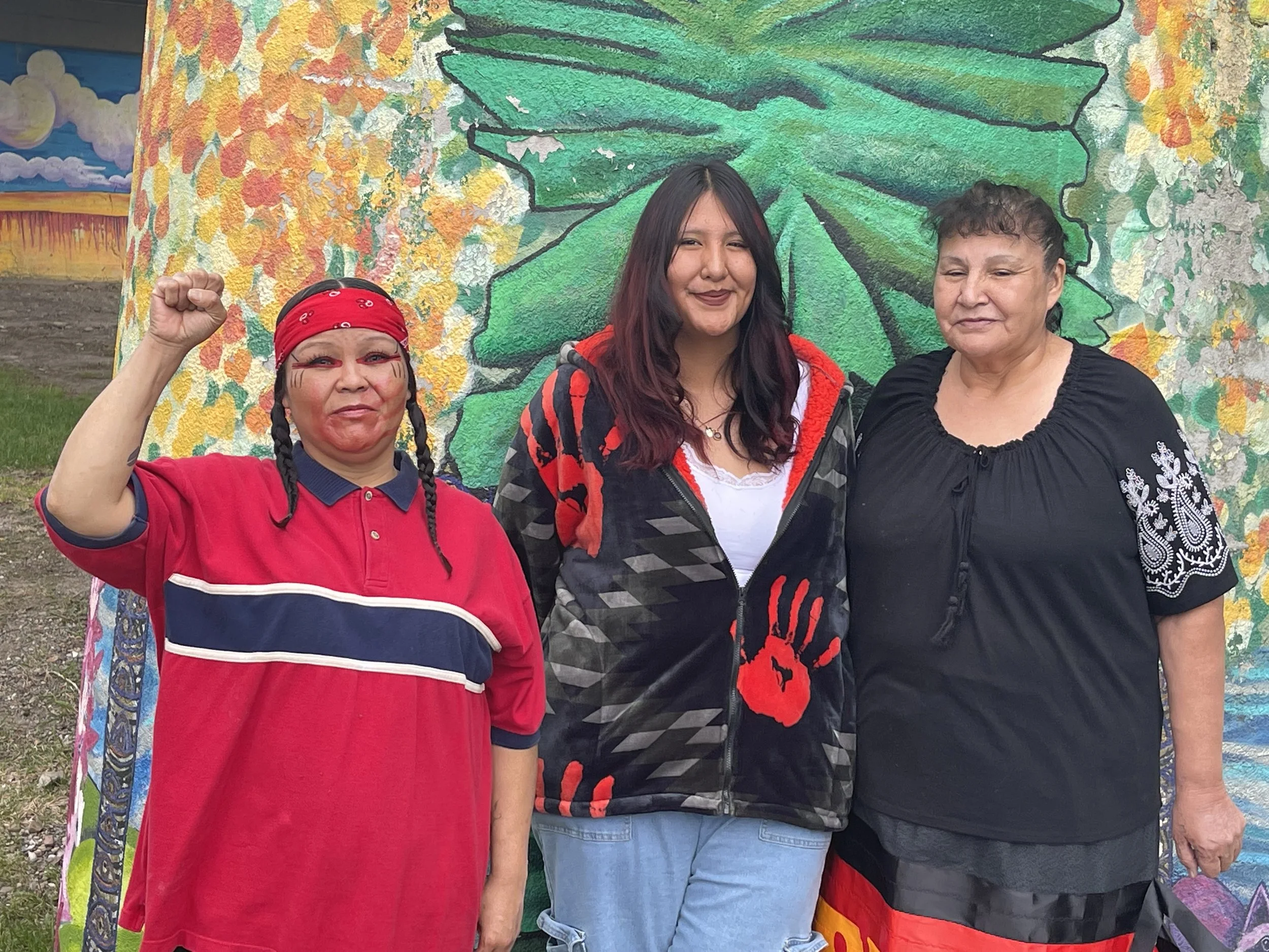 Two women are engaged in conversation at an outdoor event, one holding a clipboard and a schedule, with festival or parade attendees in the background.