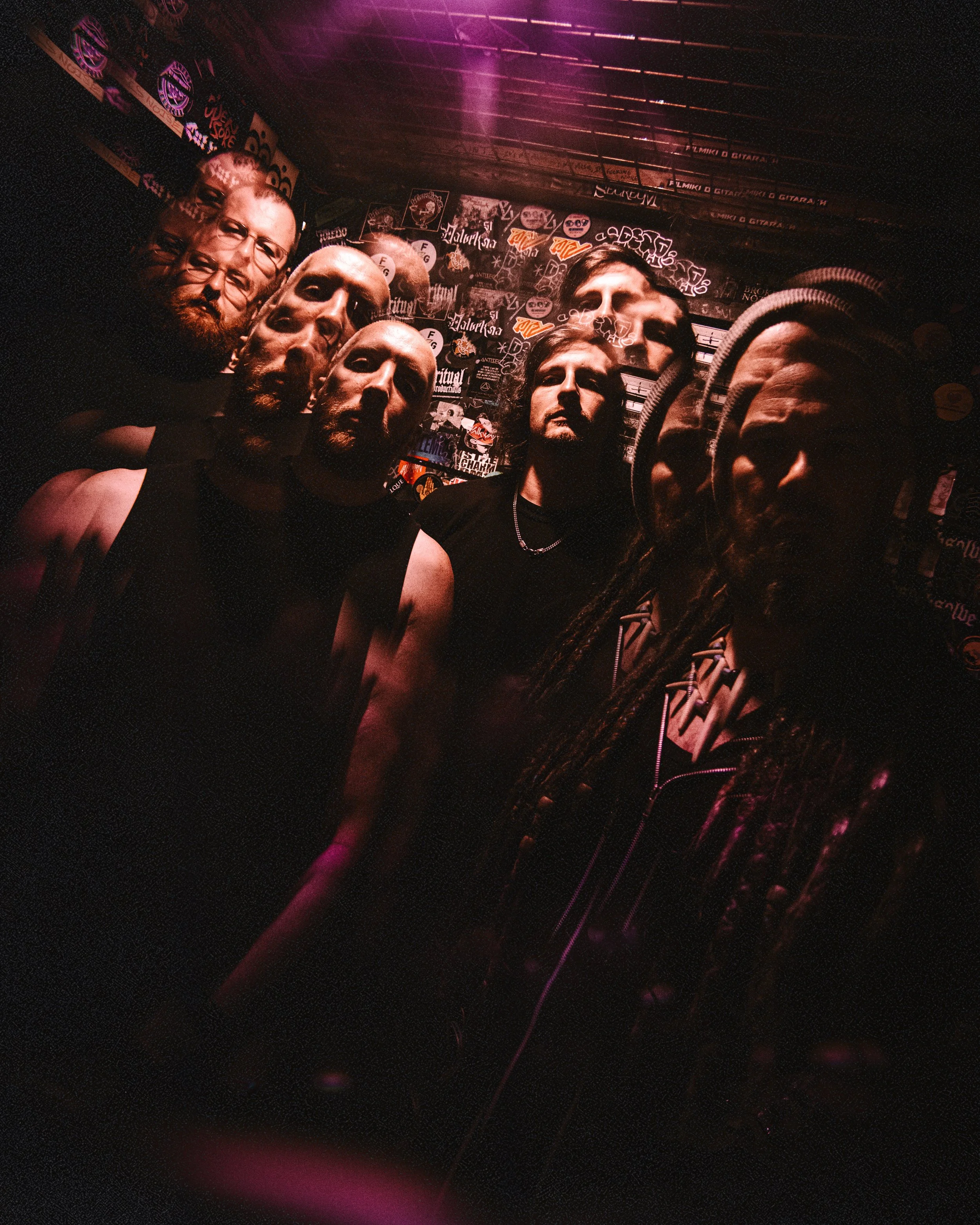 A group of seven men with beards and long hair, some wearing sleeveless shirts, posing together in a dimly lit room with graffiti and stickers on the walls.