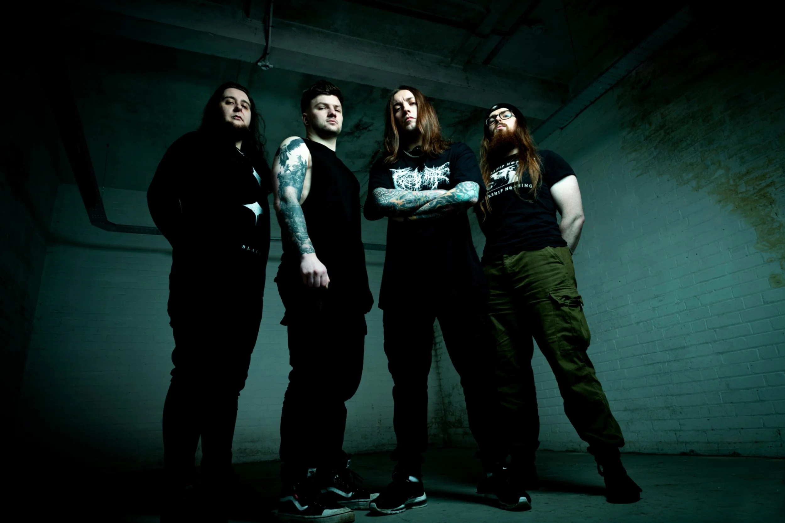 Four men with long hair and tattoos standing in a dimly lit, grungy underground parking garage with concrete walls.