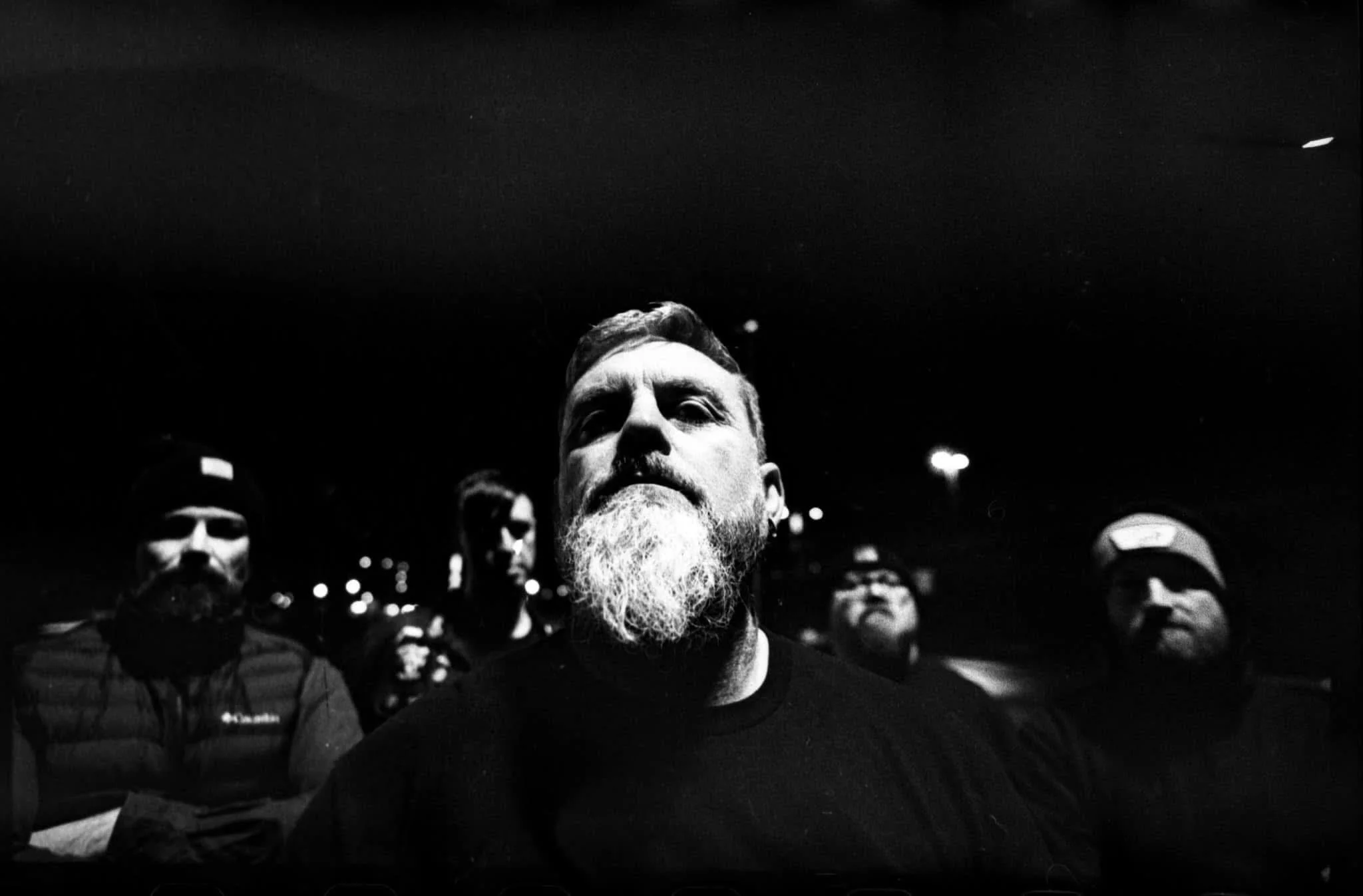 Black and white photo of five men, with the central man having a beard and serious expression, in dark clothing, in a dimly lit outdoor setting at night.