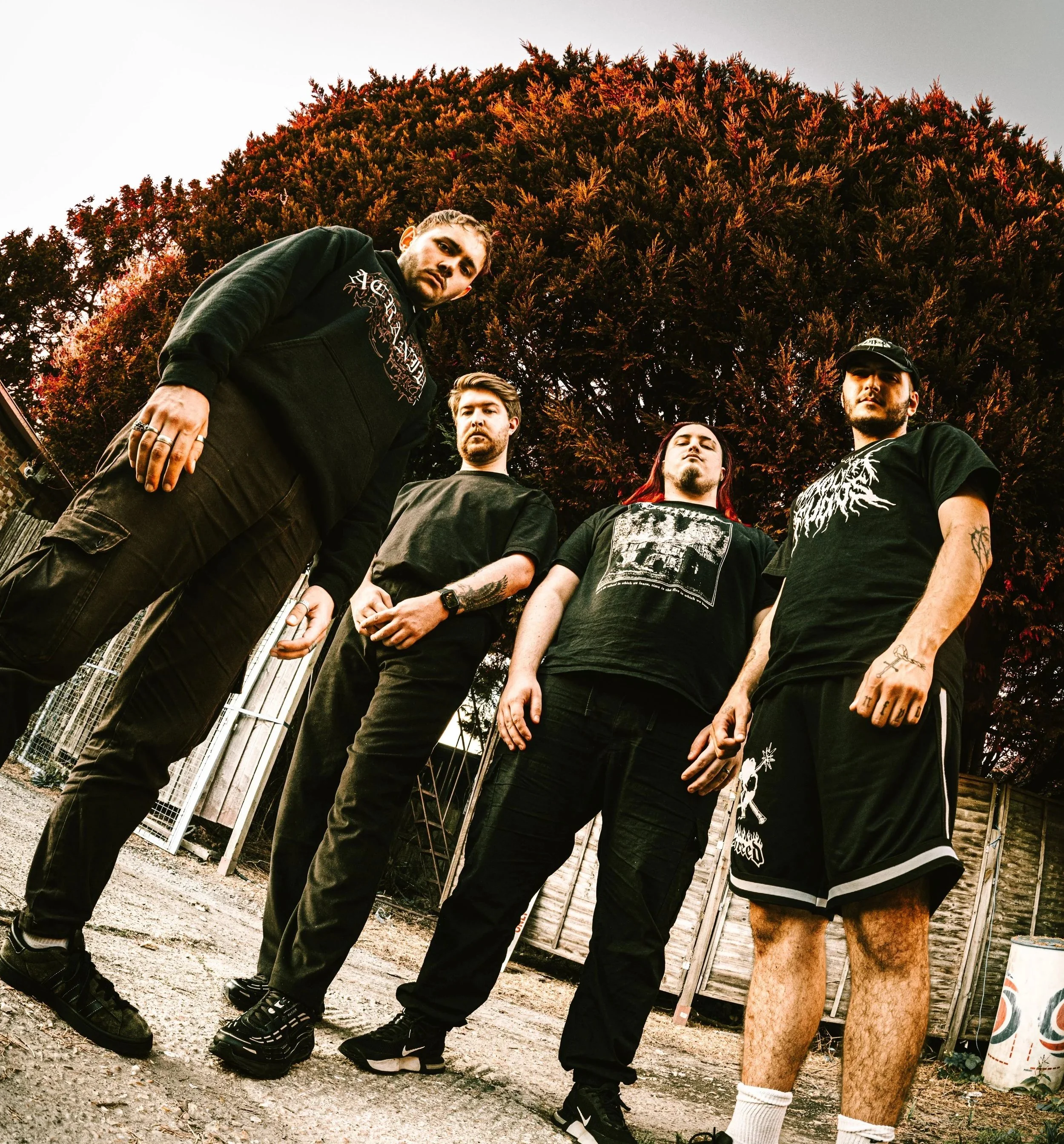 Four men standing outdoors with a large tree in the background. They are dressed in dark clothing, and some have tattoos. The photo is taken from a low angle.