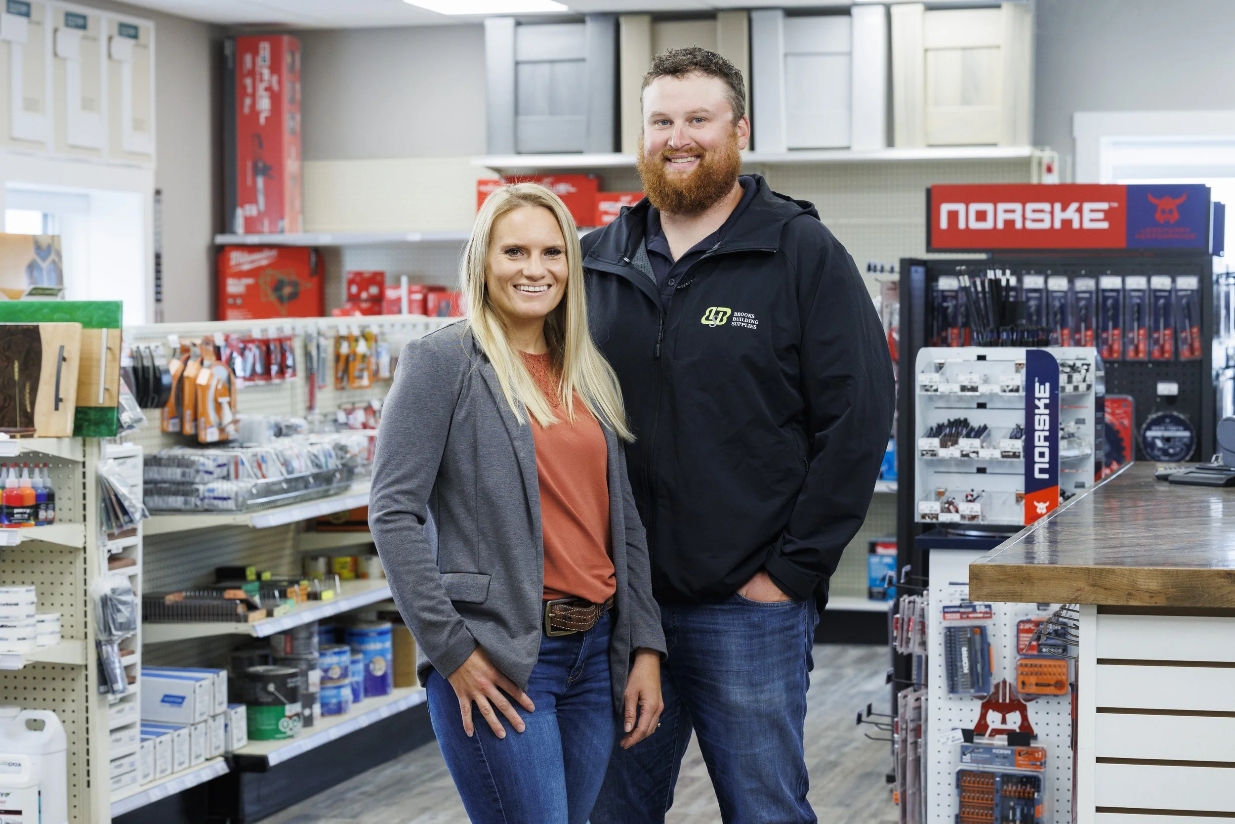 A smiling woman and man standing together inside a hardware store, surrounded by shelves stocked with tools and building supplies.