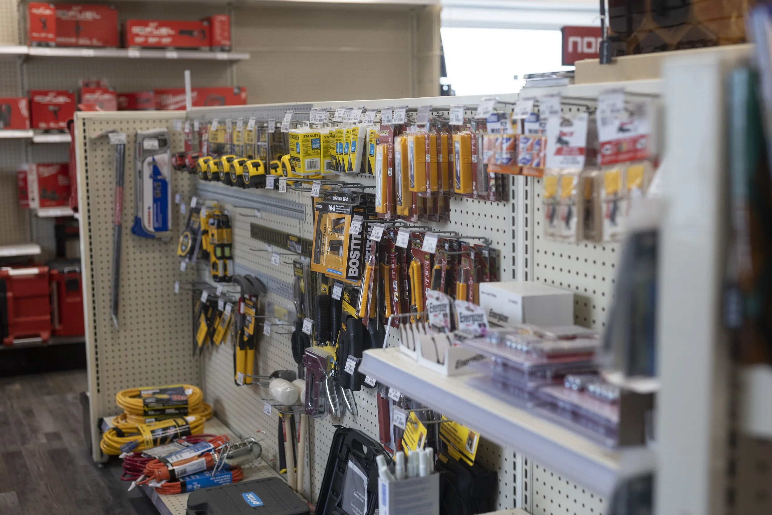 A store aisle with tools and hardware supplies displayed on pegboards, including tape measures, saws, and extension cords.