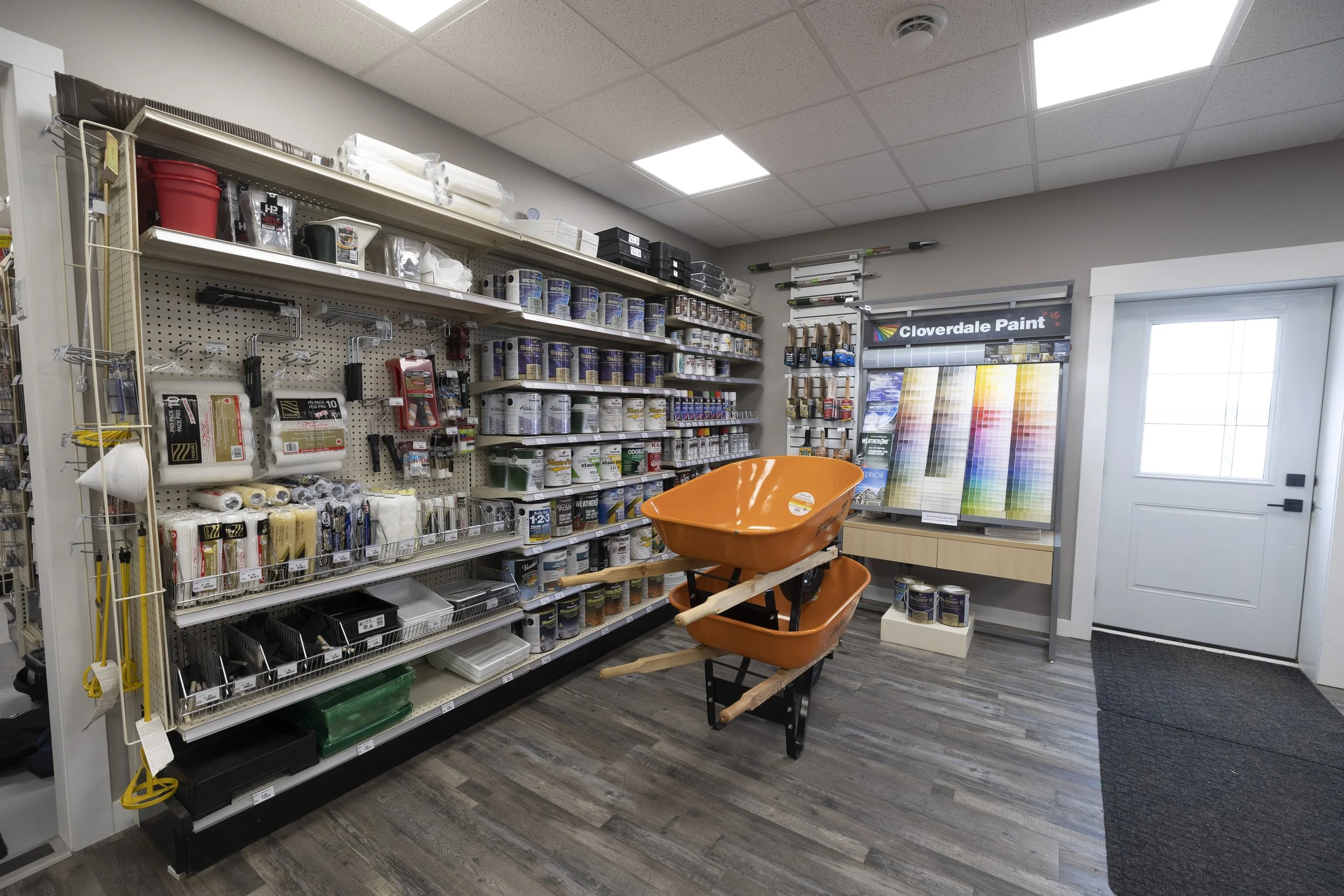 Interior of a hardware store aisle with paint supplies, tools, and a shopping cart with orange wheelbarrows.