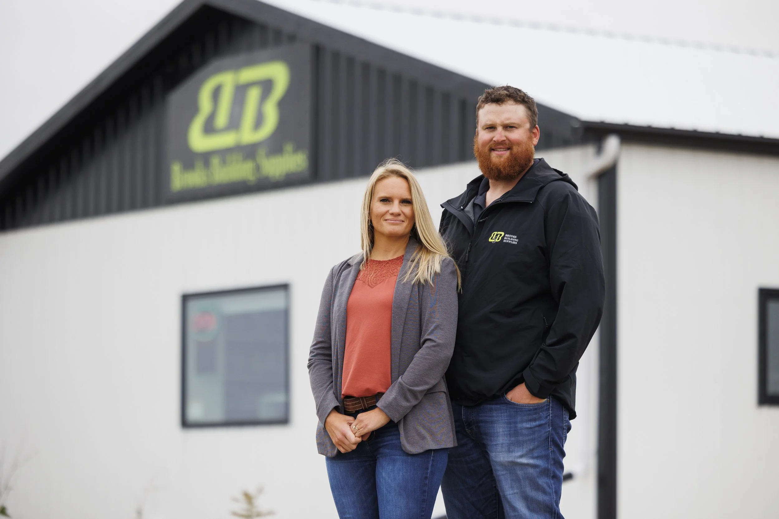 A man and a woman standing outside in front of a building with a sign that reads 'Brooks Building Supplies' in bright green and black.