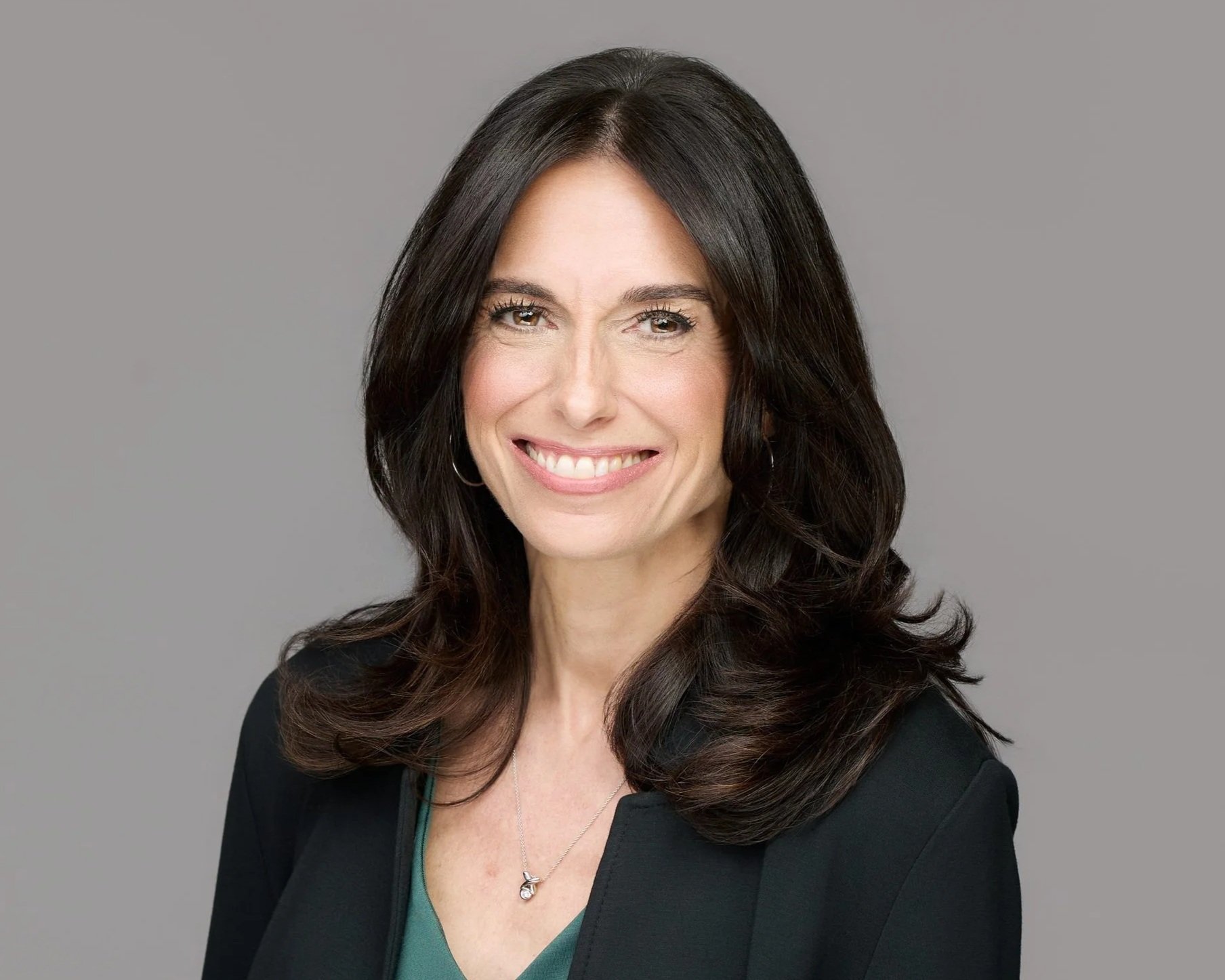 Portrait of a woman with long dark hair, smiling, wearing a black blazer and a green top, against a plain gray background.