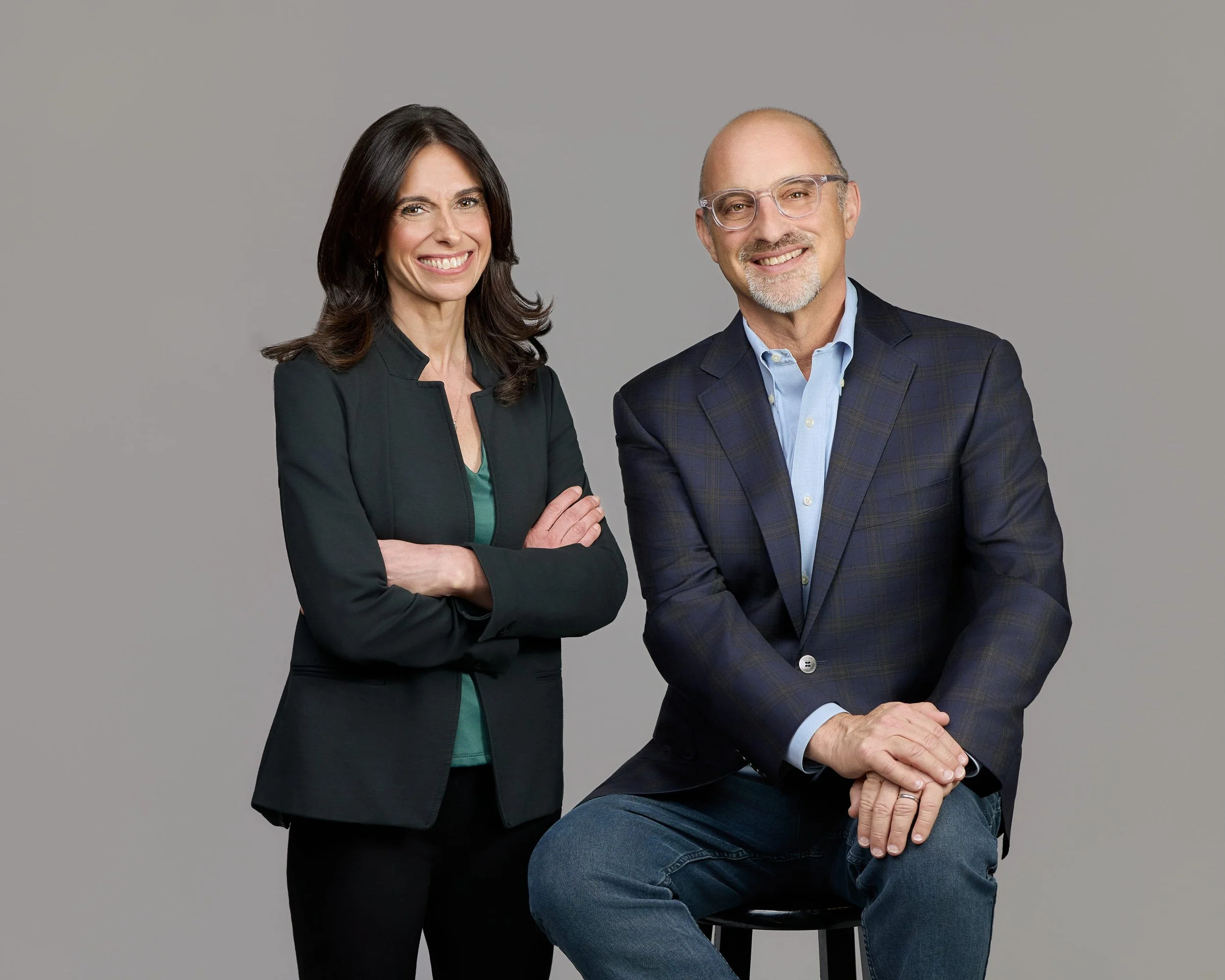 A woman with dark hair wearing a black blazer and teal top standing next to a man with glasses, a beard, wearing a plaid blazer and light blue shirt, sitting on a stool against a plain gray background.